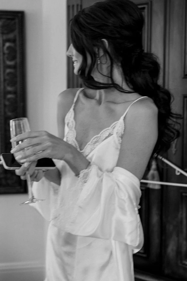A woman in a satin slip holding a glass of champagne and a smartphone, smiling and looking to her left. She has dark wavy hair and is wearing earrings, standing indoors with a wooden door behind her.