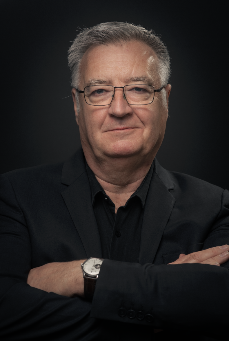 Un homme d'âge moyen portant des lunettes, un costume noir et une montre, avec une expression confiante et croisant les bras, fond noir.