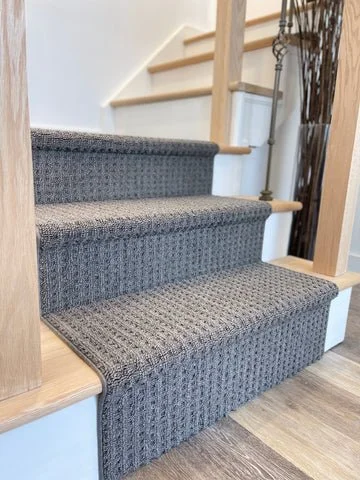 Carpeted staircase with three steps, partially enclosed by wooden staircase and railing, with hardwood flooring at the bottom.