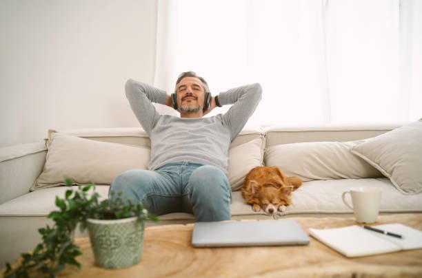 A man relaxes on a sofa with headphones, eyes closed, smiling, with a dog lying beside him, in a bright room with a coffee table, a plant, a mug, and a laptop.