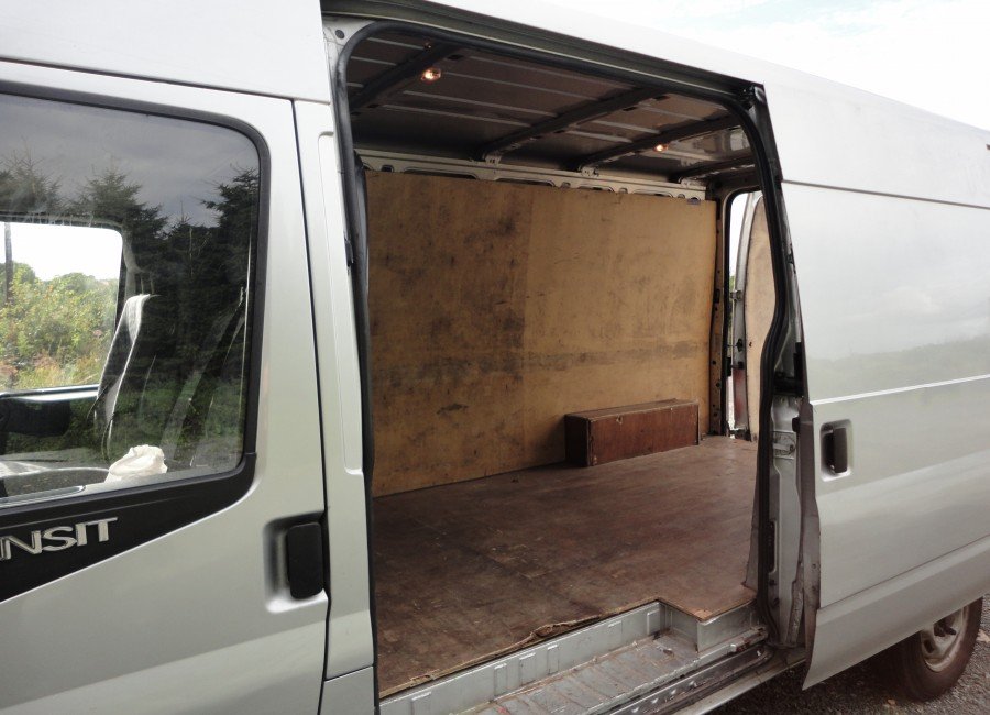 The image shows the side view of a white Ford Transit cargo van with the sliding door open, revealing an empty wooden-floored cargo space with a plywood wall and a small wooden box inside.