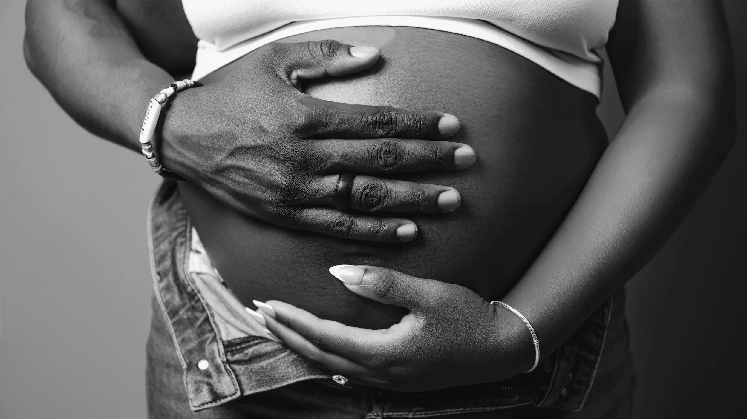 Close-up of a pregnant woman with dark skin, gently holding her belly with both hands. The photo is in black and white, showing her wearing a bracelet and a ring.