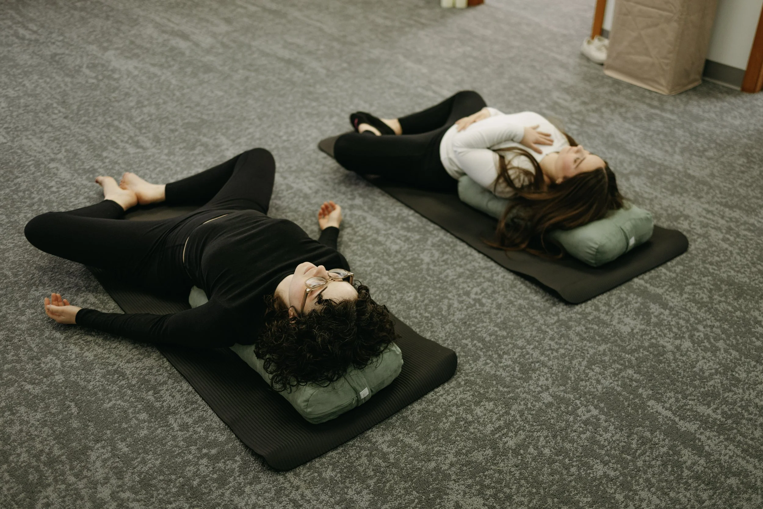 Group of people practicing yoga outdoors at sunrise, facing away, on mats in a grassy field surrounded by trees.