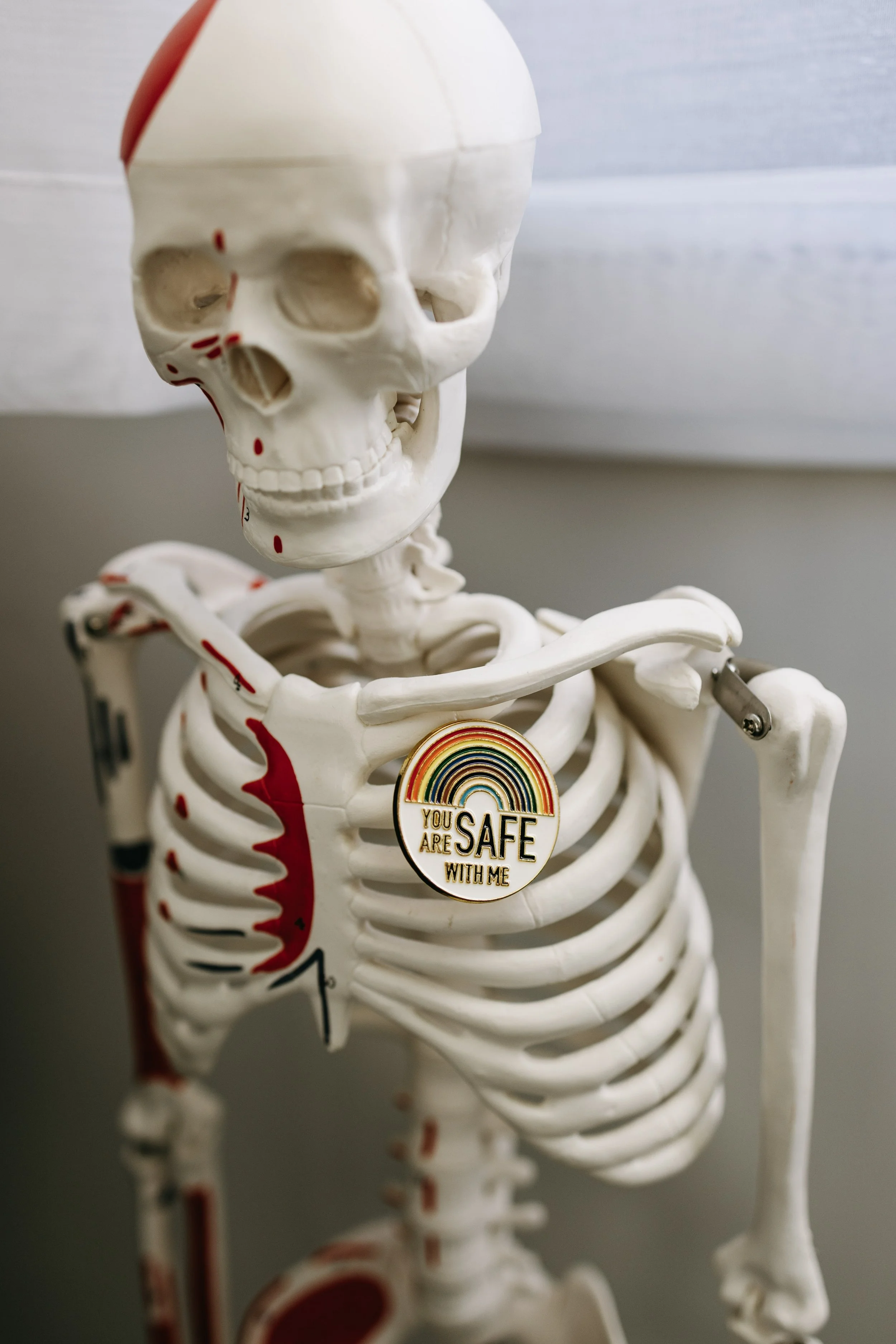 A rainbow pride flag with an additional white stripe, purple circle, black stripe, and brown stripe flying on a flagpole against a partly cloudy sky with leafless tree branches.