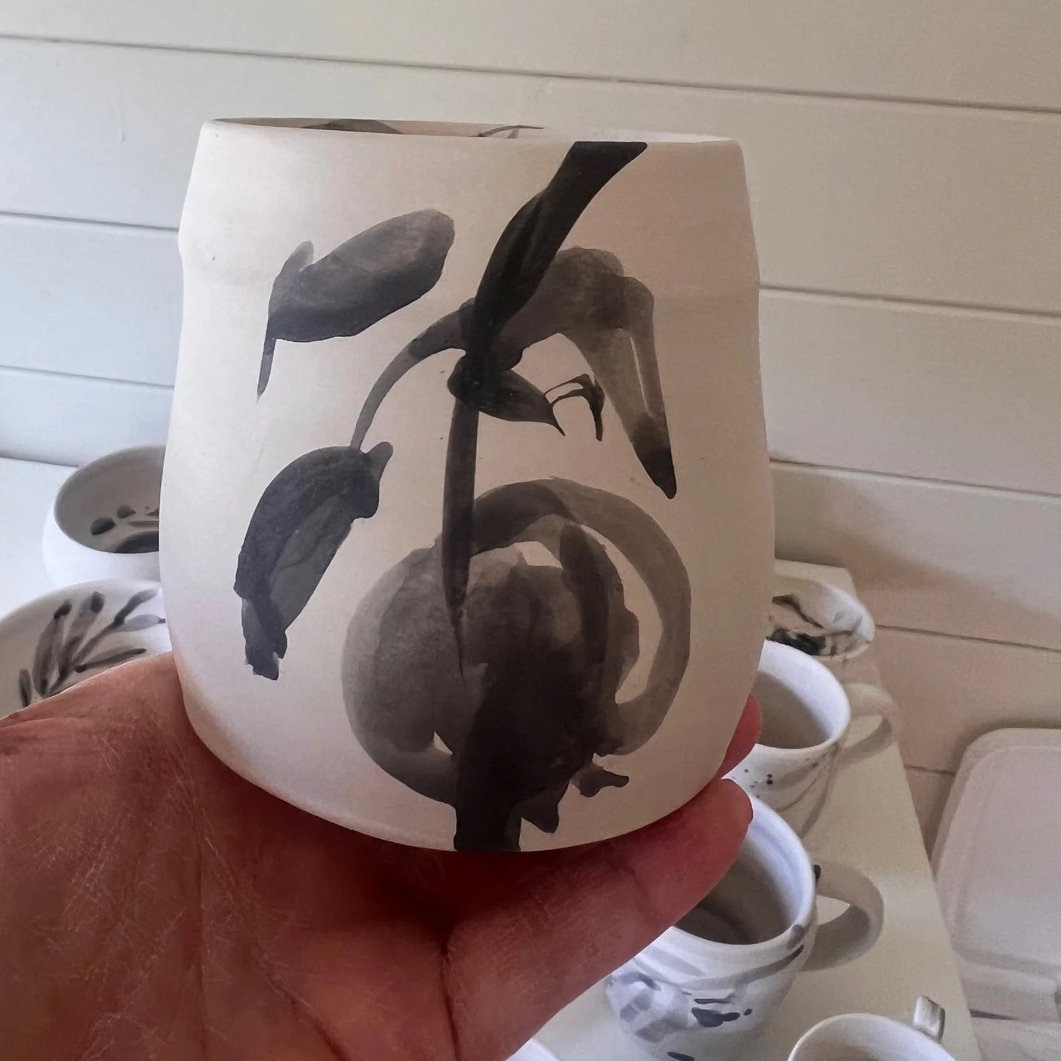 A white ceramic mug with a black and white artistic print of a plant with leaves and a curved stem, held in a person's hand. Several similar mugs with black and white plant designs are visible in the background.