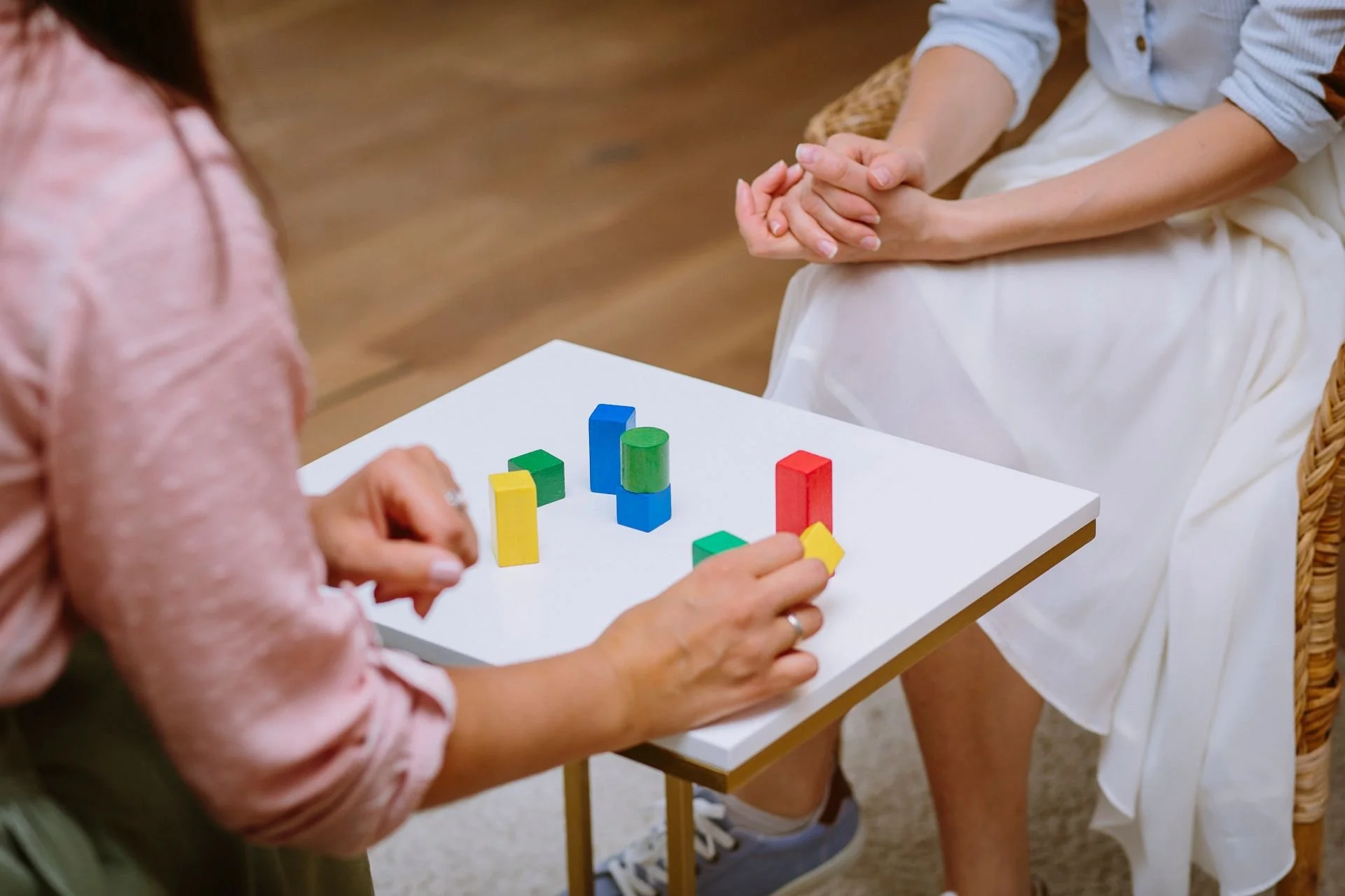 Ein weißer Tisch mit bunten Holzbausteinen, die von zwei Personen gespielt werden. Eine Person trägt eine rosa Bluse und die andere trägt ein weißes Kleid. Der Raum hat einen Holzboden.