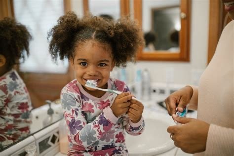 Young girl brushing her teeth in a bathroom, smiling at the camera, with a woman holding a toothbrush in the background.