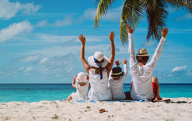 Family sitting on the beach under palm trees, enjoying the ocean view with arms raised in the air, wearing sun hats and casual summer clothes.