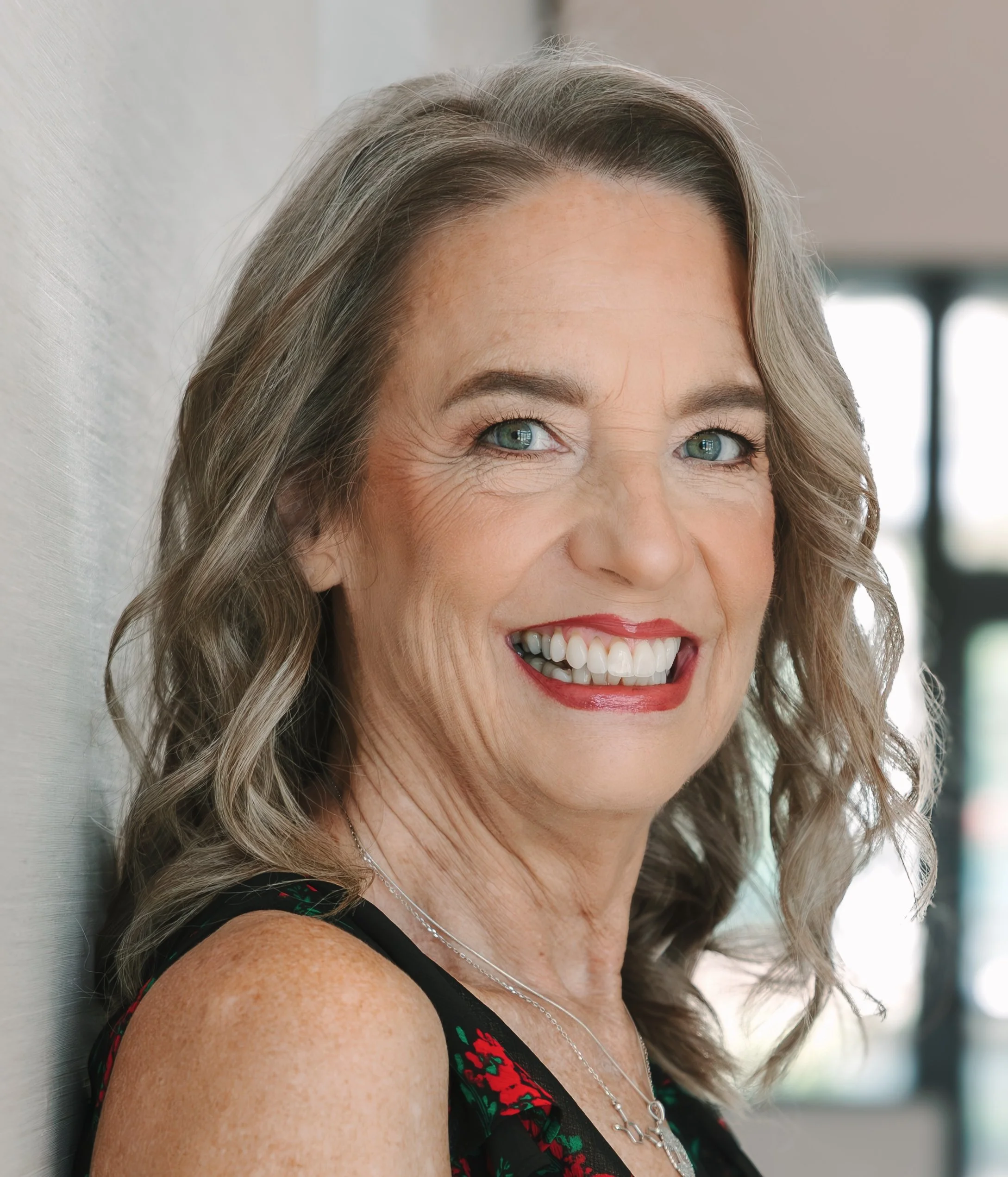 Reverend Sarah, wedding officiant, smiling in a black dress with red floral pattern, standing indoors near a wall with bright background.