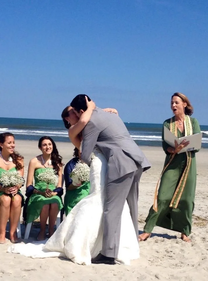 A wedding ceremony on the beach with a couple kissing, three bridesmaids holding bouquets, and an officiant reading from a book.
