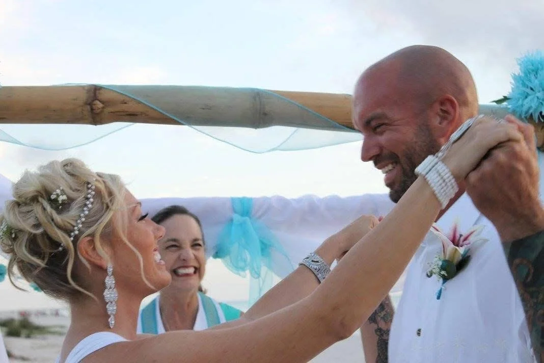 A couple smiling and sharing an embrace during their wedding ceremony on the beach in Sarasota County, Florida, with a Rev. Sarah in the background smiling.