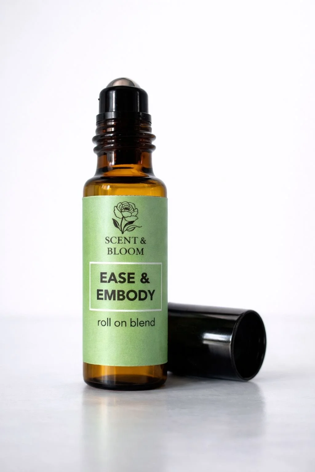 A small amber glass bottle with a black rollerball top and a black cap lying beside it. The bottle has a green label with a flower illustration and the text 'SCENT & BLOOM' and 'EASE & EMBODY roll on blend'.