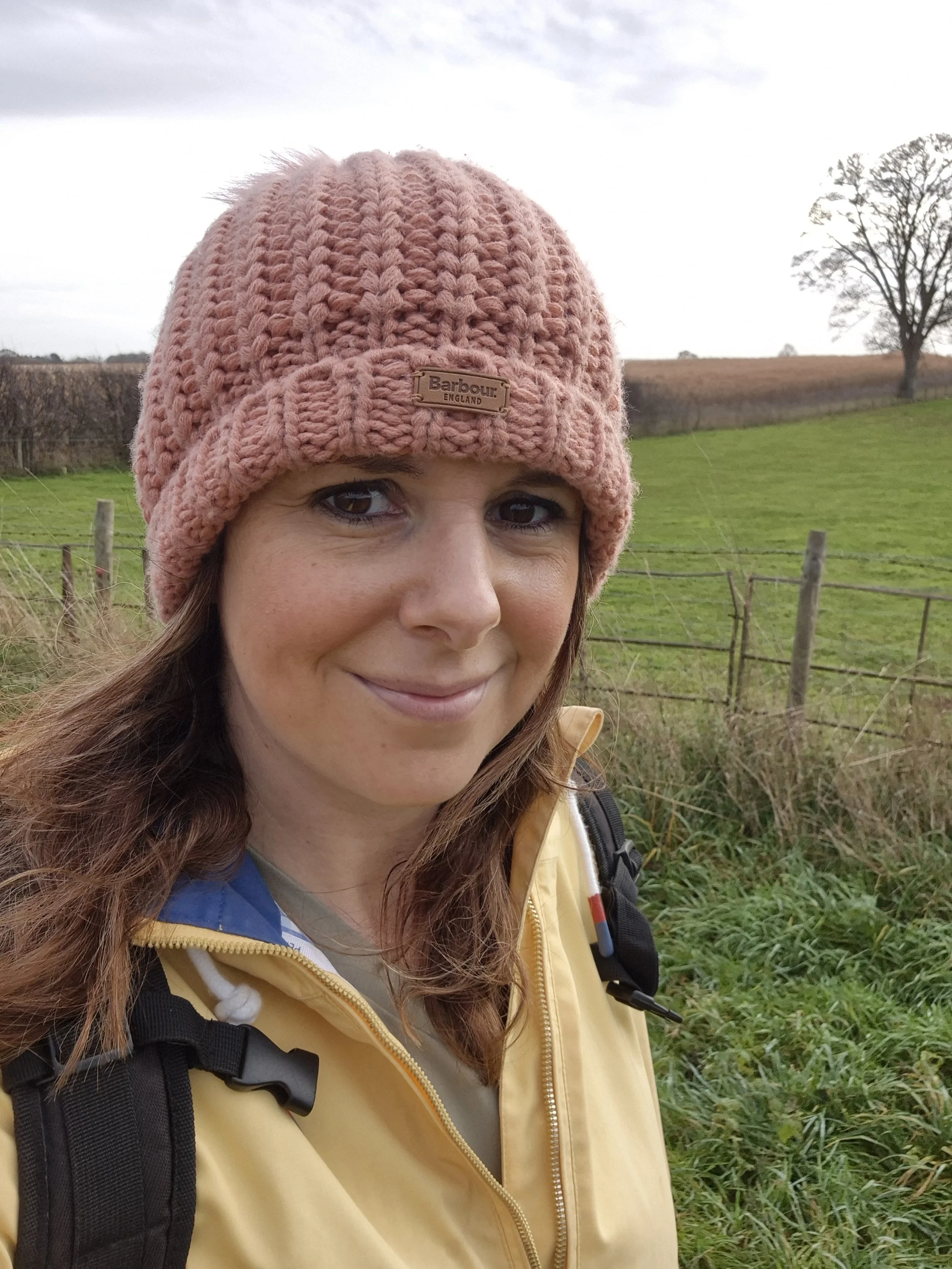 An autistic aromatherapist wearing a pink knit beanie, yellow jacket, and backpack standing outdoors in a grassy field with a fence and trees in the background, during daytime.