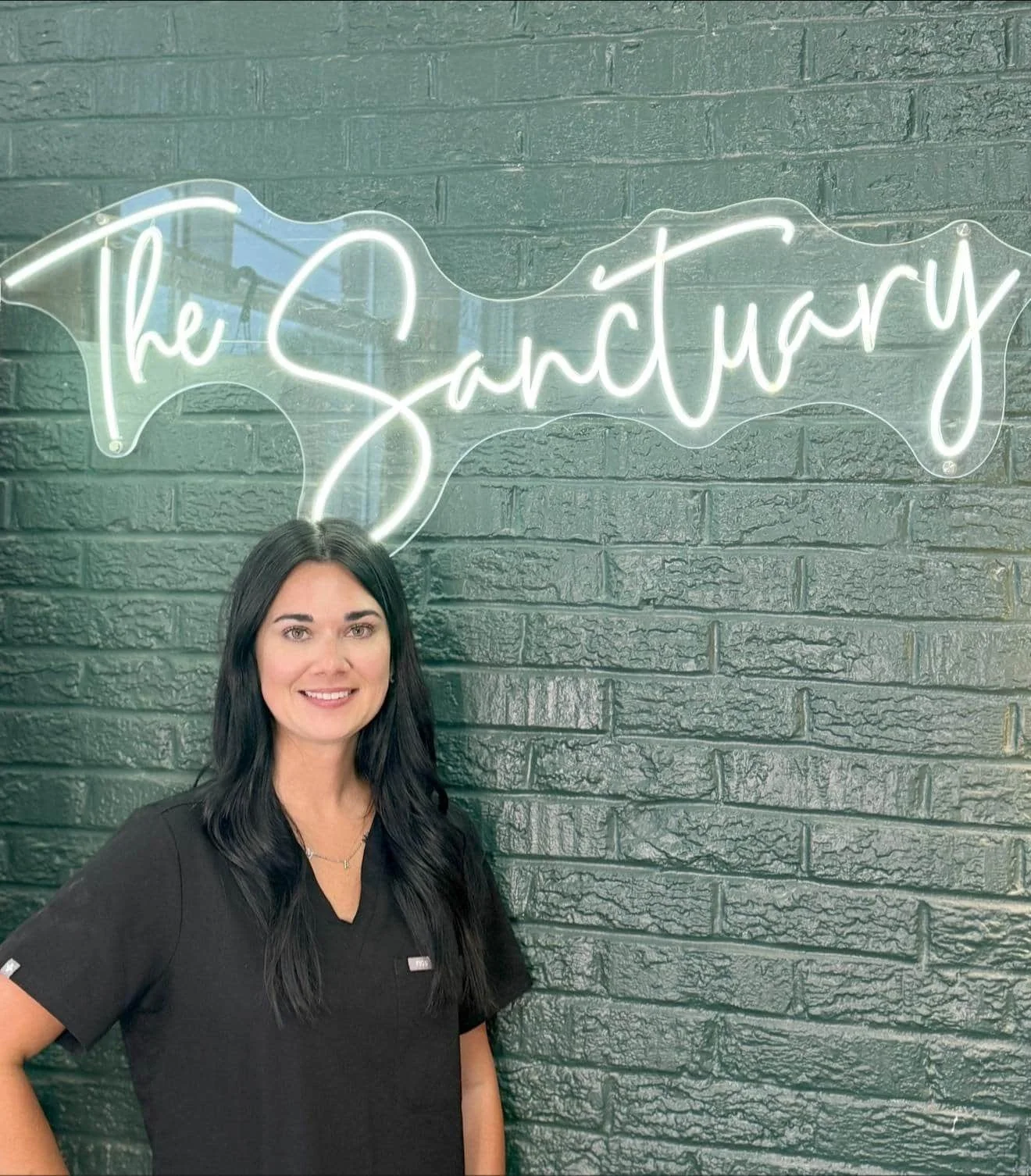 Taylor, a licensed esthetician, standing in front of The Sanctuary Spas neon sign. She is your estie bestie.