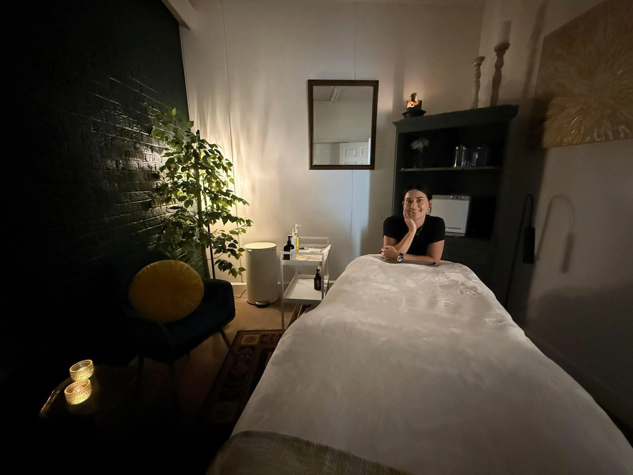 A licensed esthetician, Taylor sitting at the head of a massage table in a dimly lit room, smiling. The room has a black brick wall, a green plant, a small white table with bottles, a mirror on the wall, and candles on a side table. your estie bestie