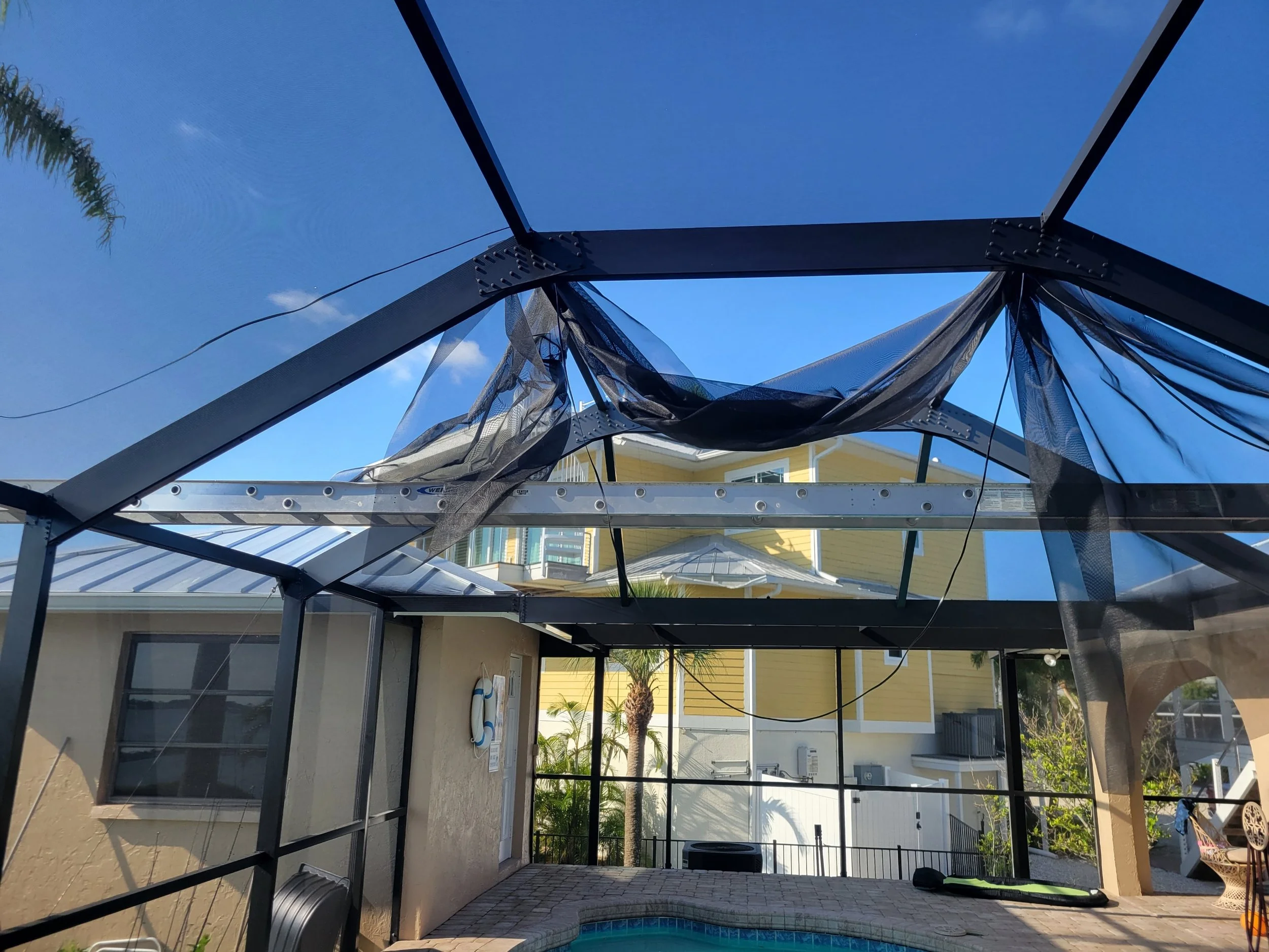 Damaged pool enclosure with fallen screen mesh and bent frame, in a backyard with a yellow house in the background.