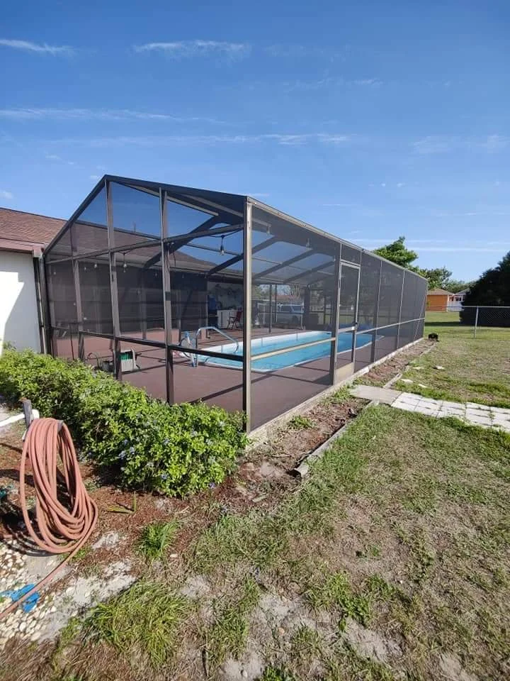 An outdoor swimming pool enclosed by a screened structure in a backyard, with a concrete patio and some greenery outside.