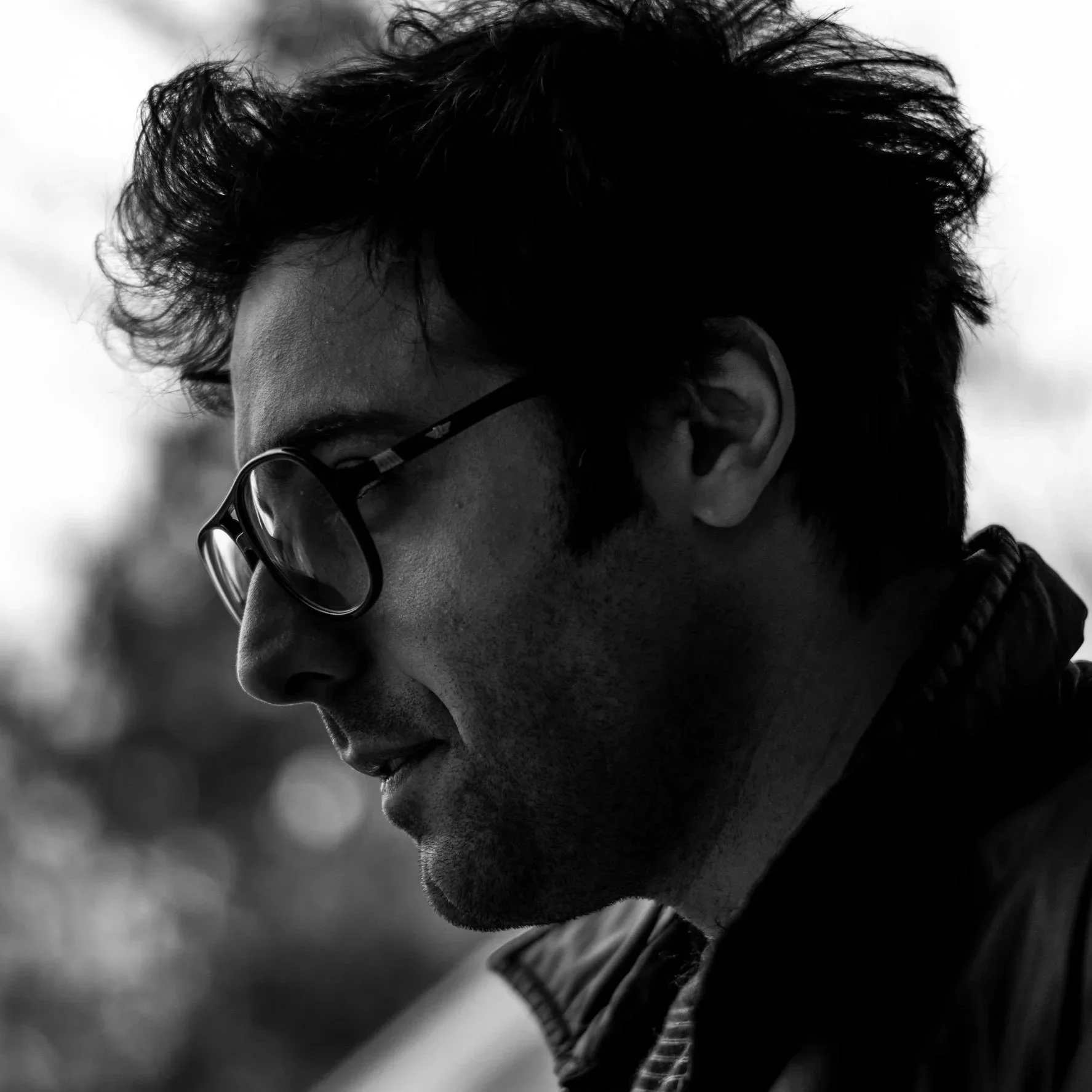 Black and white close-up photo of a young man with curly hair, wearing glasses, looking down