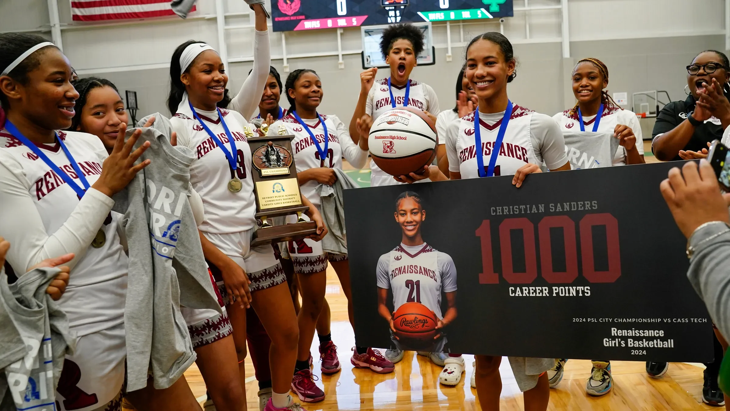 Group of young girls in basketball uniforms celebrating a victory, holding a trophy and a basketball, with a large sign proclaiming Christian Sanders' achievement of 1,000 career points in girls' basketball.