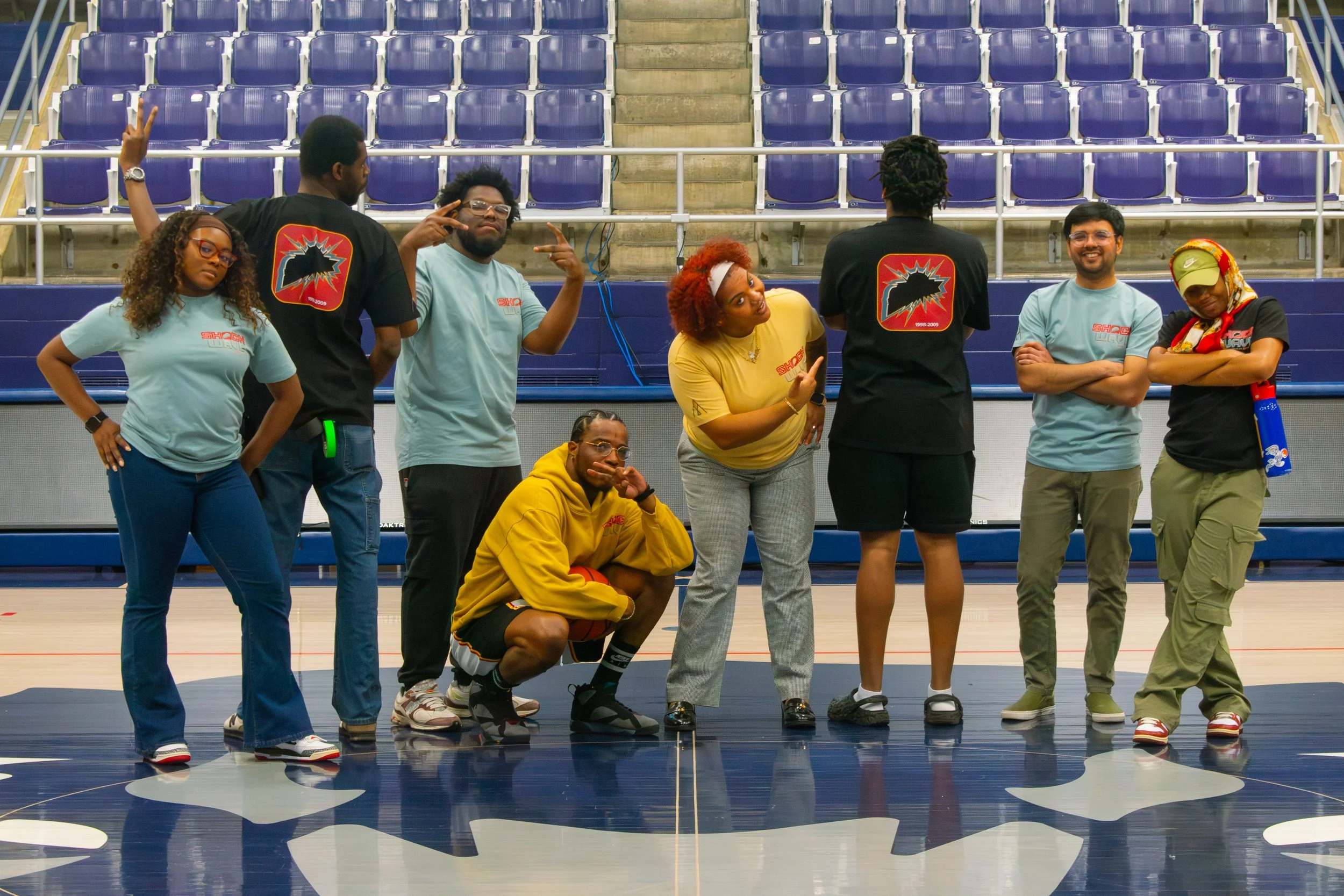 Group of six people posing inside a sports arena, some standing and one crouching, with empty blue and purple stadium seats in the background. They are wearing casual and sportswear, with some making peace signs and other playful gestures.