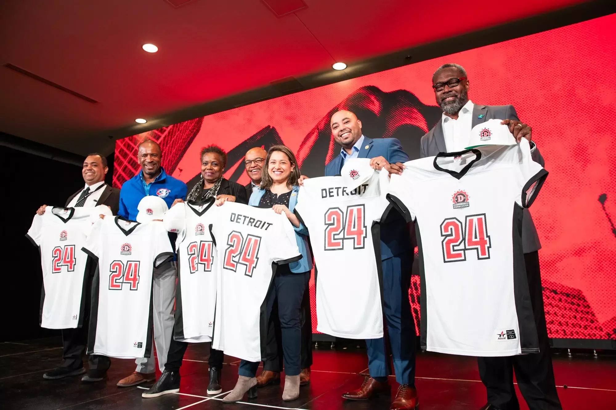 Group of diverse people holding Detroit basketball jerseys with the number 24, standing on stage during a formal event.