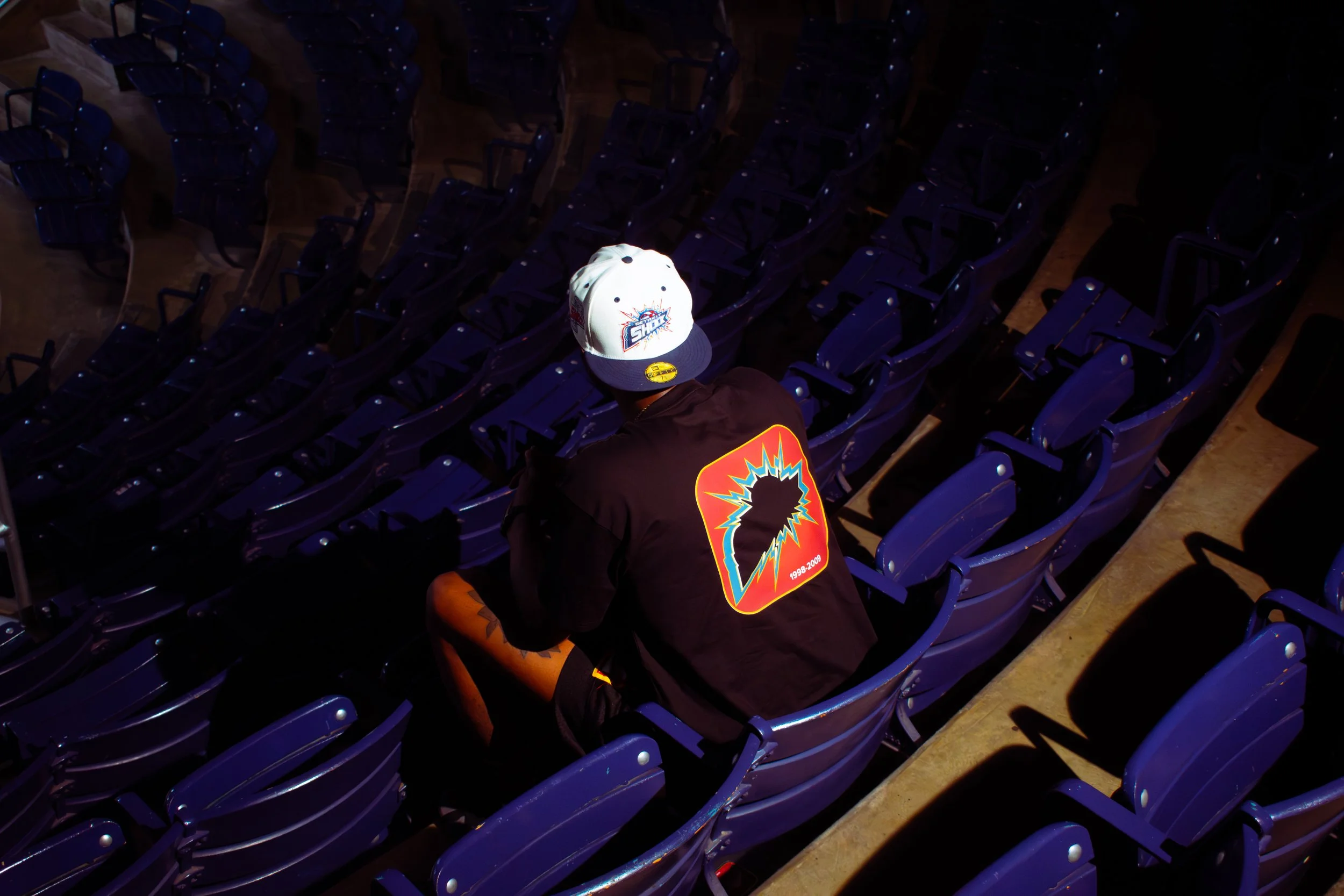A person wearing a cap and a black t-shirt with vibrant graphics is sitting alone in a large stadium with many empty purple chairs. The image is taken from above.