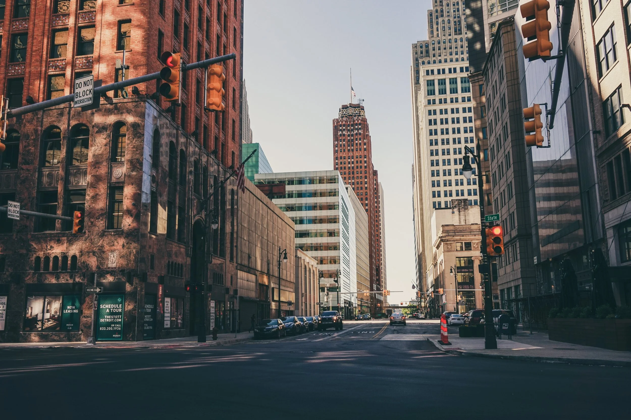 City street scene with tall buildings on both sides, traffic lights, parked cars, and a partly cloudy sky.