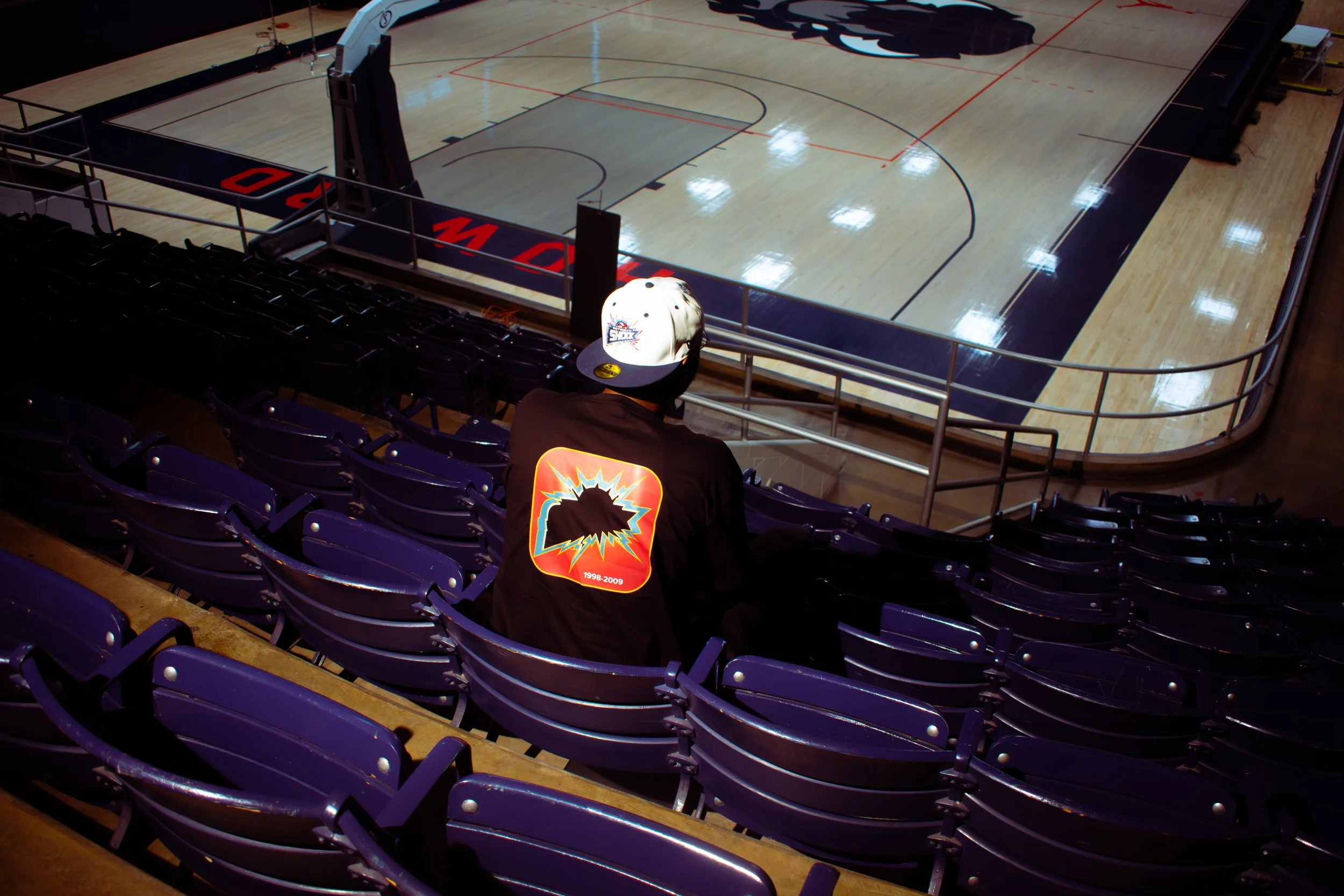 An empty basketball court viewed from the stands with a person sitting in the front row wearing a cap and a black shirt with a colorful graphic on the back, in an arena with purple seats.