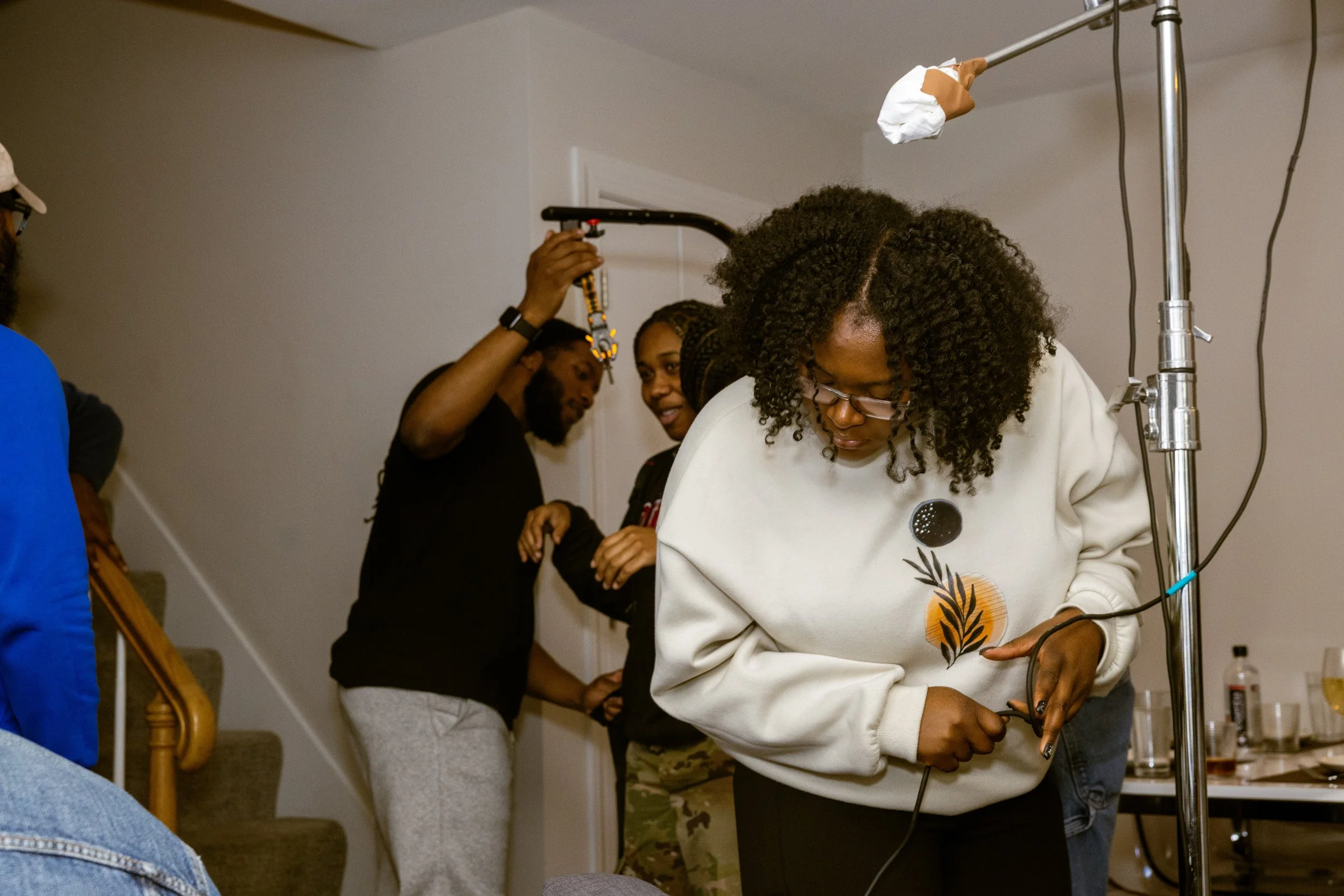 Several people gathered indoors, one woman with curly hair and glasses working on equipment, others watching or talking.
