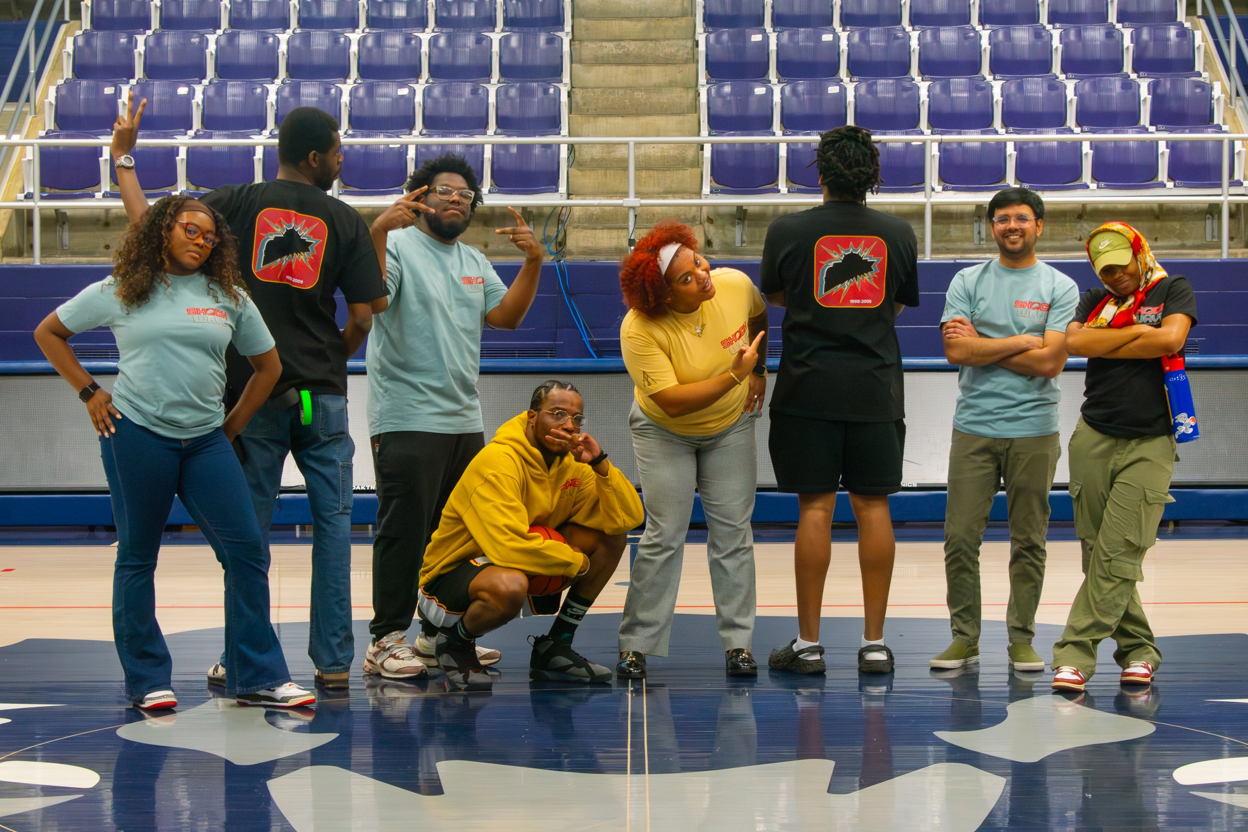 Group of seven young adults posing on an indoor basketball court, some making playful gestures, with empty blue stadium seats in the background.