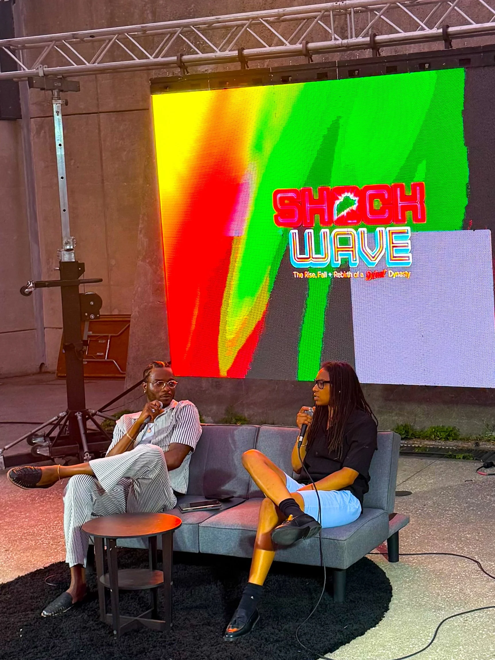 Two people sitting on a gray sofa with a black rug underneath, engaged in a conversation. Behind them, a large digital screen displays a colorful graphic with the words 'Shock Wave' and a subtitle about the rise, fall, and rebirth of a dynasty. One person with glasses and striped pants appears relaxed, while the other with glasses, a dark shirt, and light-colored shorts is holding a microphone, suggesting an interview or discussion.
