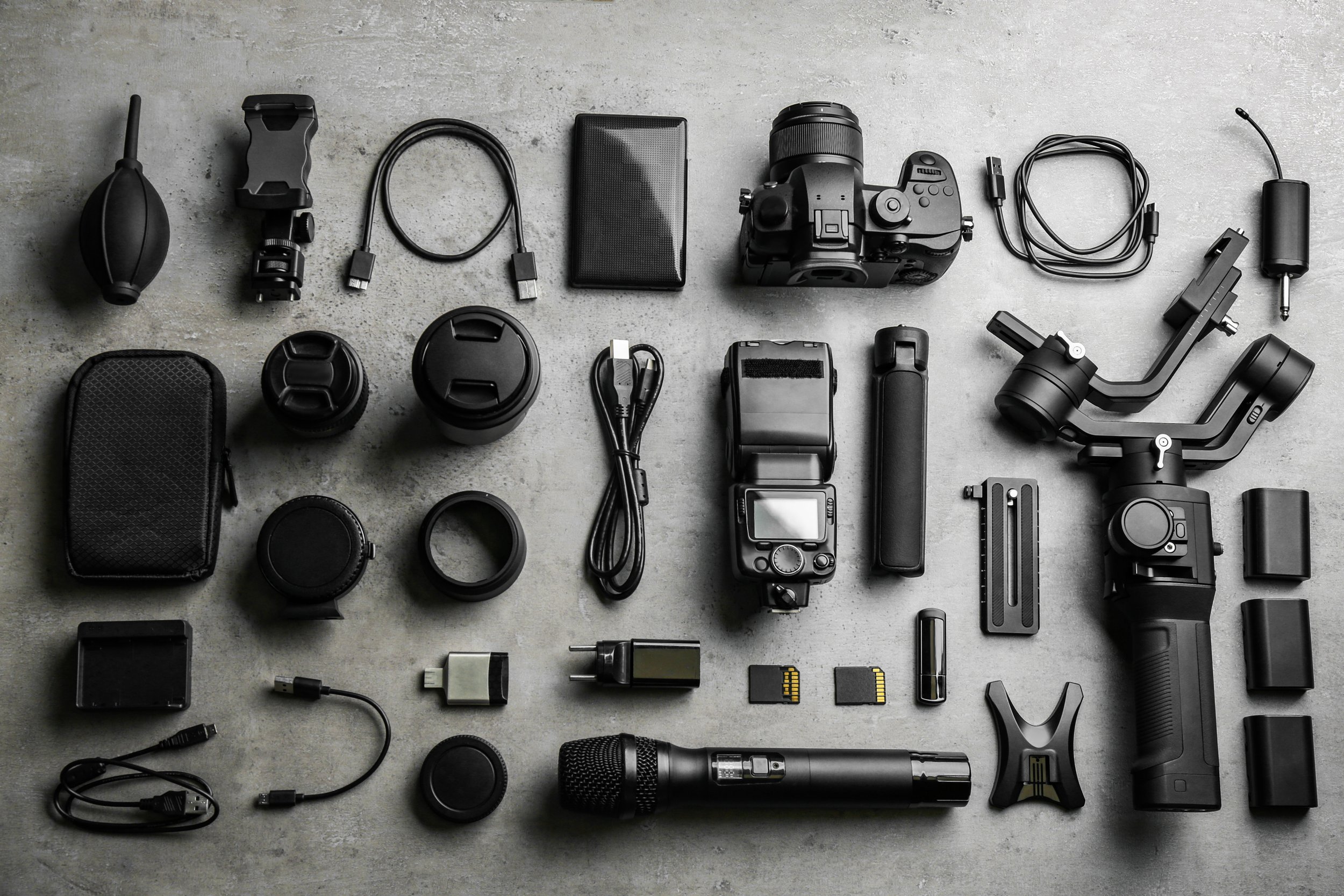A flat lay of various photography and videography equipment on a gray surface, including cameras, lenses, batteries, chargers, cables, and accessories.