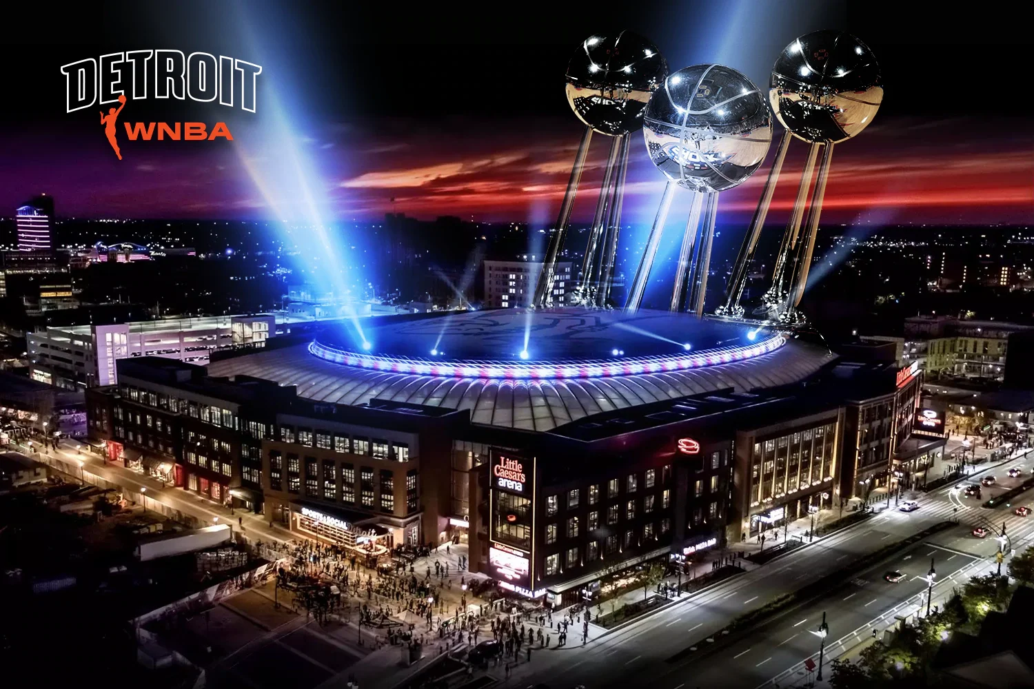 Nighttime aerial view of Little Caesars Arena in Detroit with a lit-up roof. Large, reflective trophies or sculptures are displayed on top of the building, illuminated by colorful lights. The surrounding cityscape is visible with streets and crowds outside the arena.
