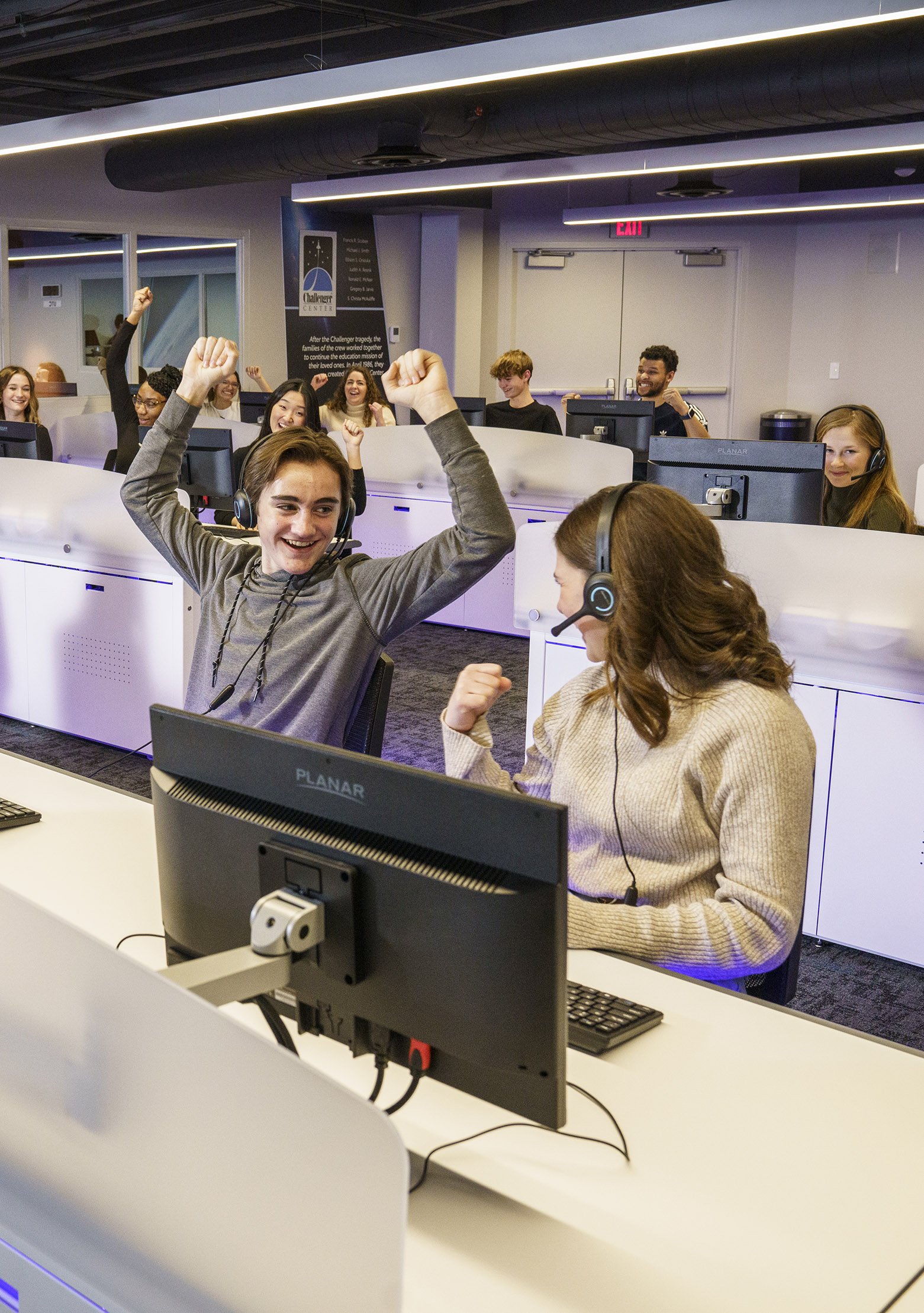 Students in the Challenger Center celebrating, with one student in the foreground raising his arms in excitement and another student smiling and gesturing with her hand, all wearing headsets and sitting at computer stations.