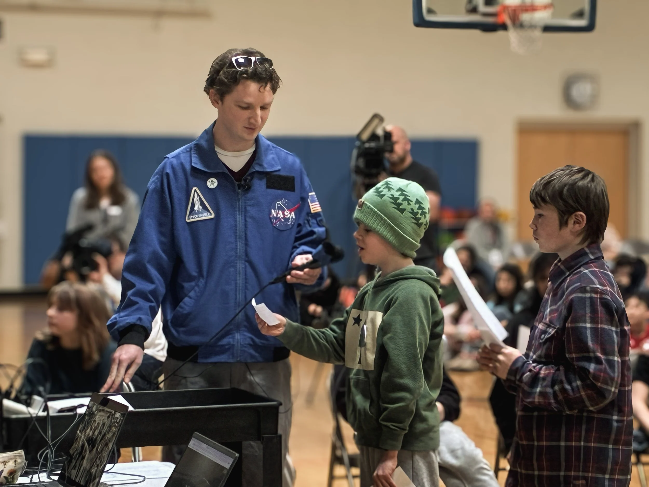 A boy in a green hoodie and a matching green beanie with tree patterns speaking to a man in a NASA jacket during a school event or presentation. The boy is holding a piece of paper, and other students and a woman are seated in the background.