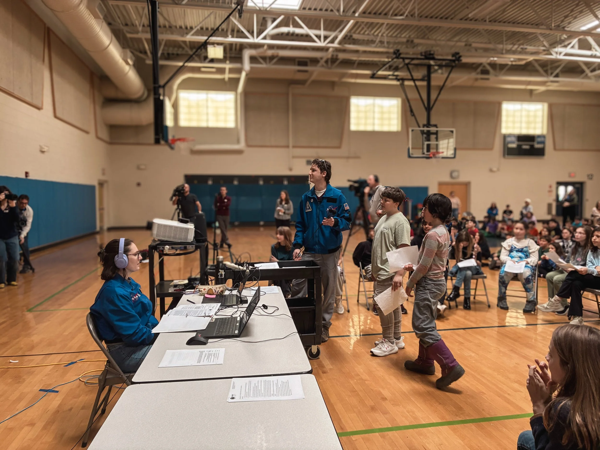 Children and adults participating in a school or community event inside a gymnasium with tables, chairs, and a basketball hoop. Several people, including a woman at a table with computers and a woman with headphones, are overseeing the event. Kids are standing in line holding papers, while others sit on chairs in the background.
