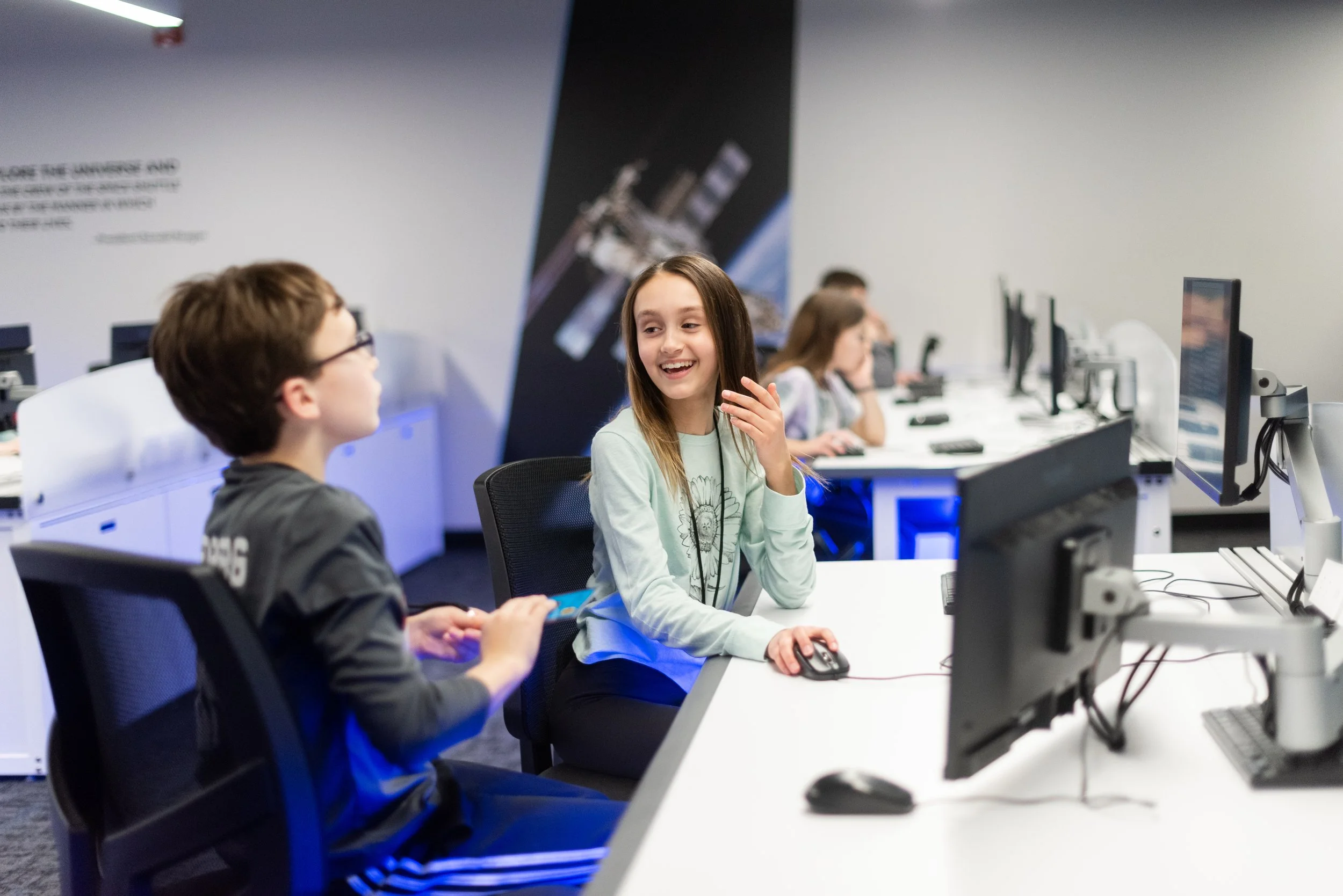 Young girl and boy talking in front of computers in a classroom or computer lab setting.