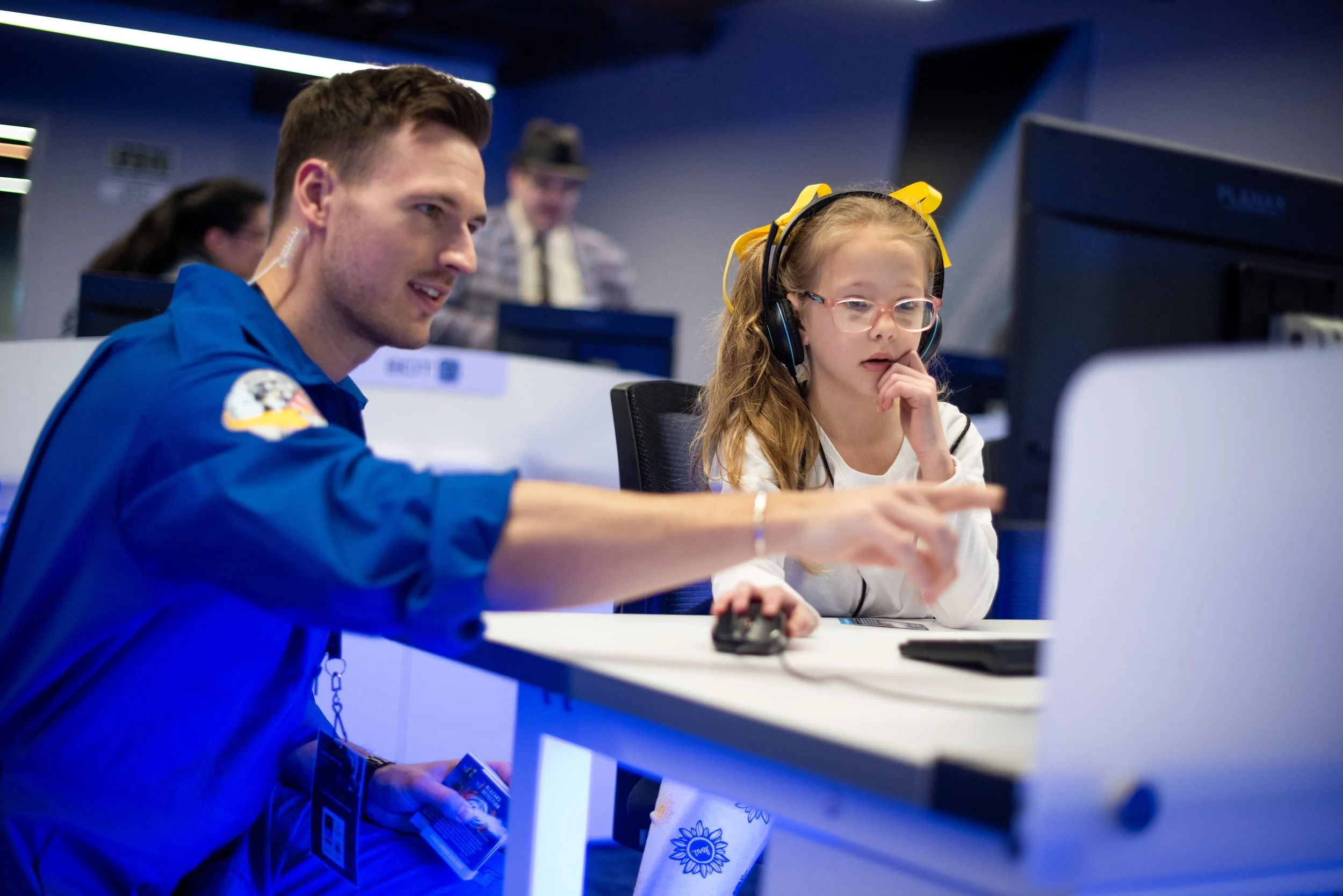 An adult instructor in a blue jumpsuit sitting with a young student wearing glasses, lookng at a computer monitor and pointing