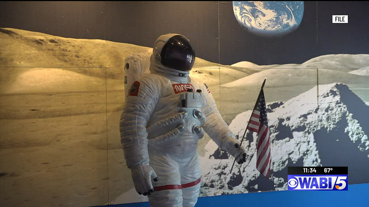 A life-sized astronaut suit holding an American flag in front of a mural of the moon's surface with Earth visible in the sky.