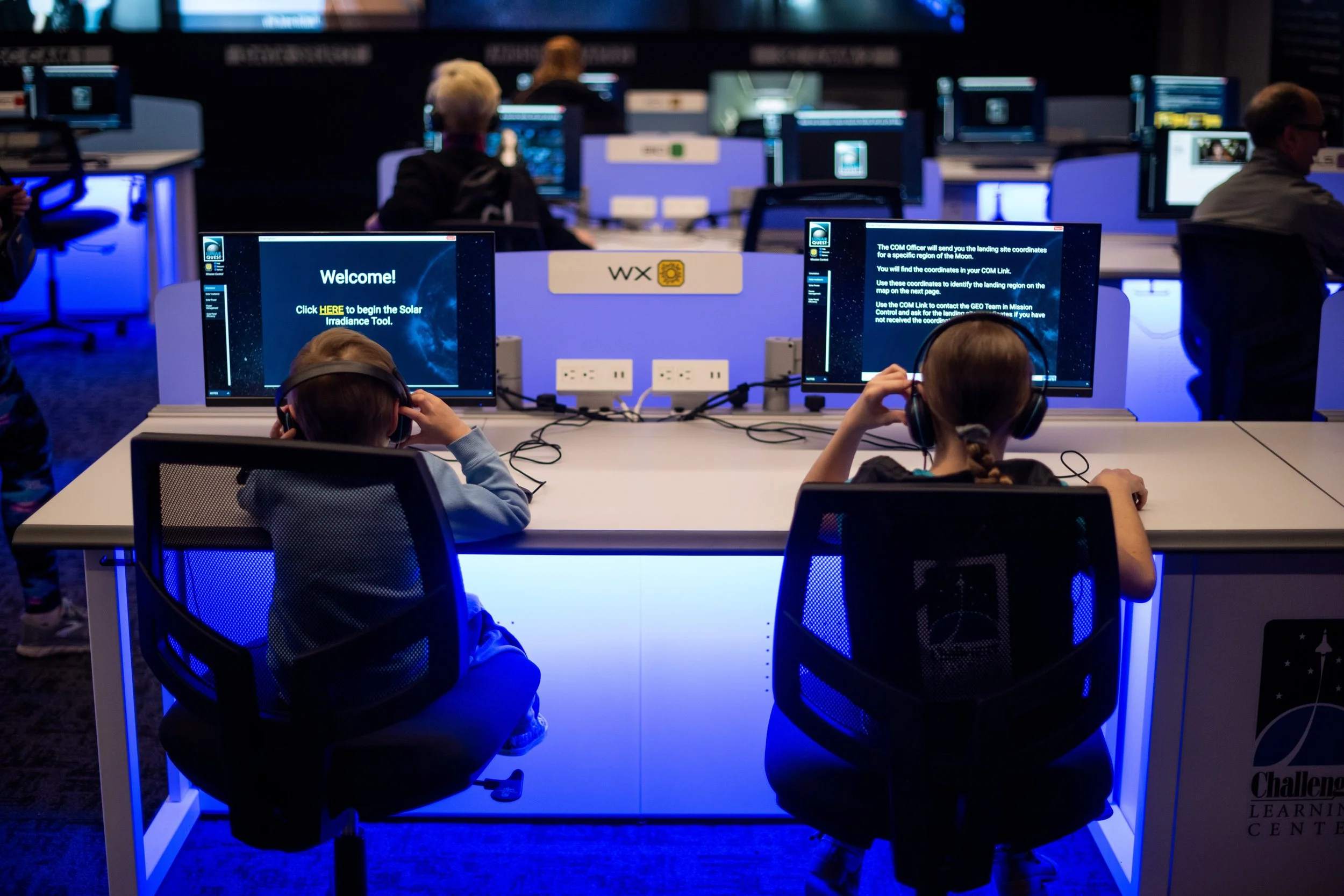 Two children sitting at a desk in front of computer monitors, wearing headphones, in a room with multiple computer workstations, with a focus on the screen that displays a welcome message for a solar irradiance tool, illuminated with blue lighting.
