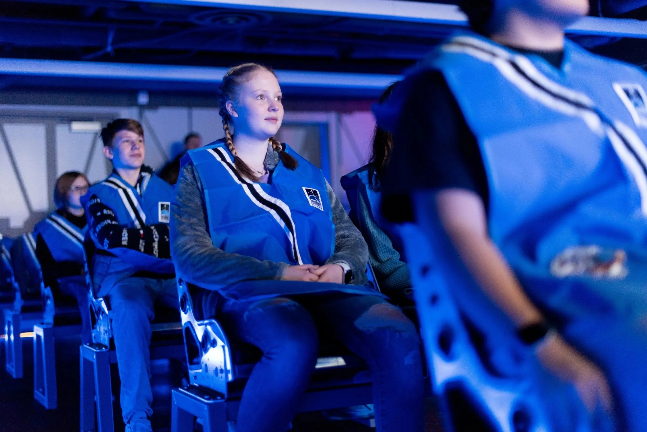 Children sitting in the Challenger transport area wearing blue astronaut vests, paying attention to a presentation in a dark, futuristic environment.