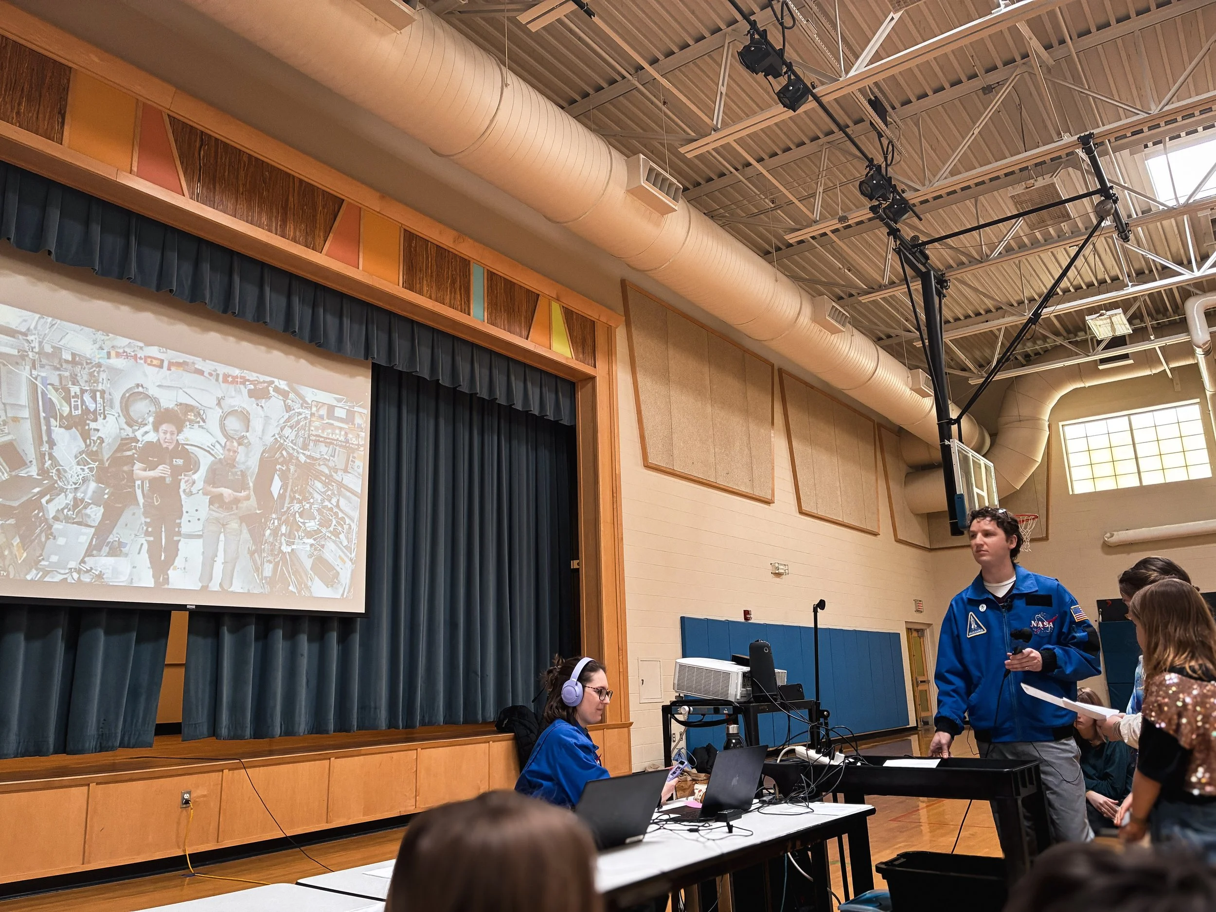 A classroom or auditorium with a stage, a large projection screen displaying a photo of people in a space or laboratory setting, and individuals participating in a presentation or activity. A woman with headphones sits at a desk with laptops, and a man in a blue NASA jacket is standing near a girl, possibly giving a presentation.