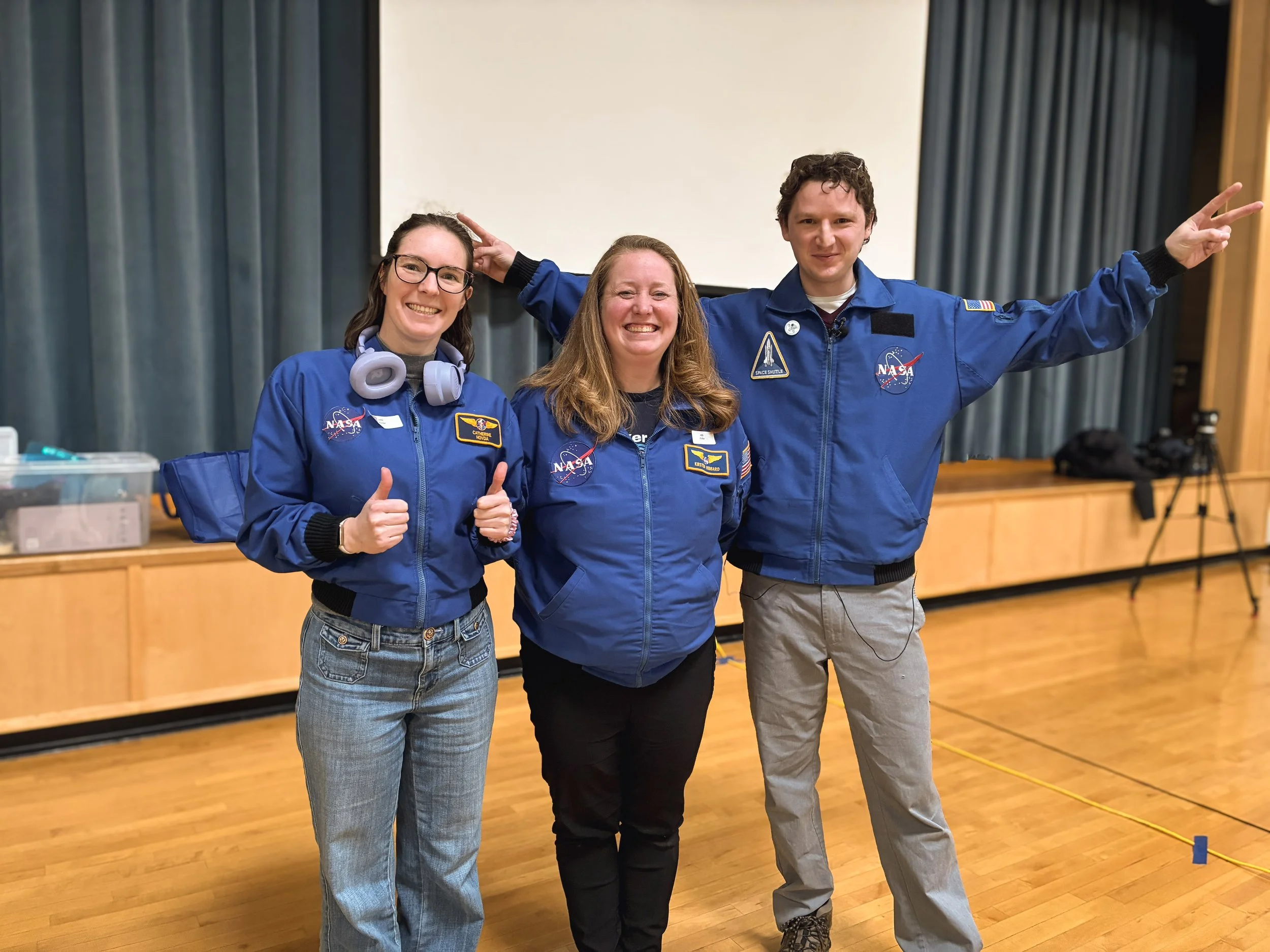 Three people in NASA astronaut jackets posing together in a room, smiling, with one giving a thumbs-up and another making peace signs.
