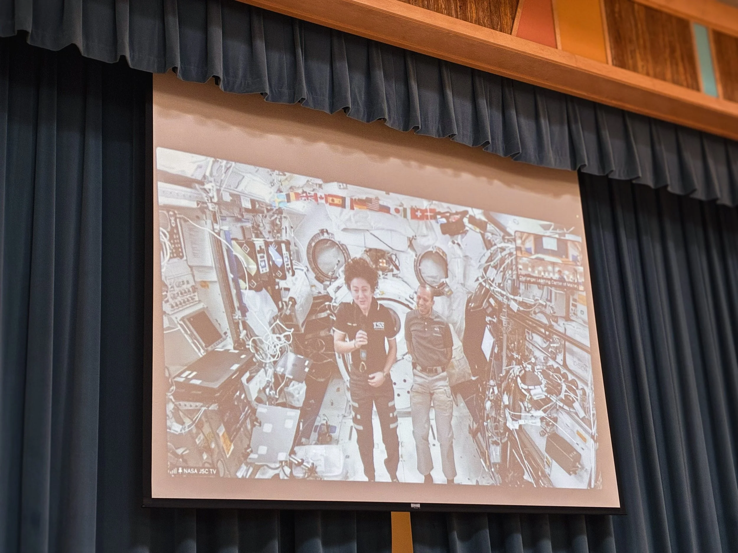 Astronauts inside the International Space Station surrounded by equipment and electronics, with one astronaut holding a microphone and both smiling.
