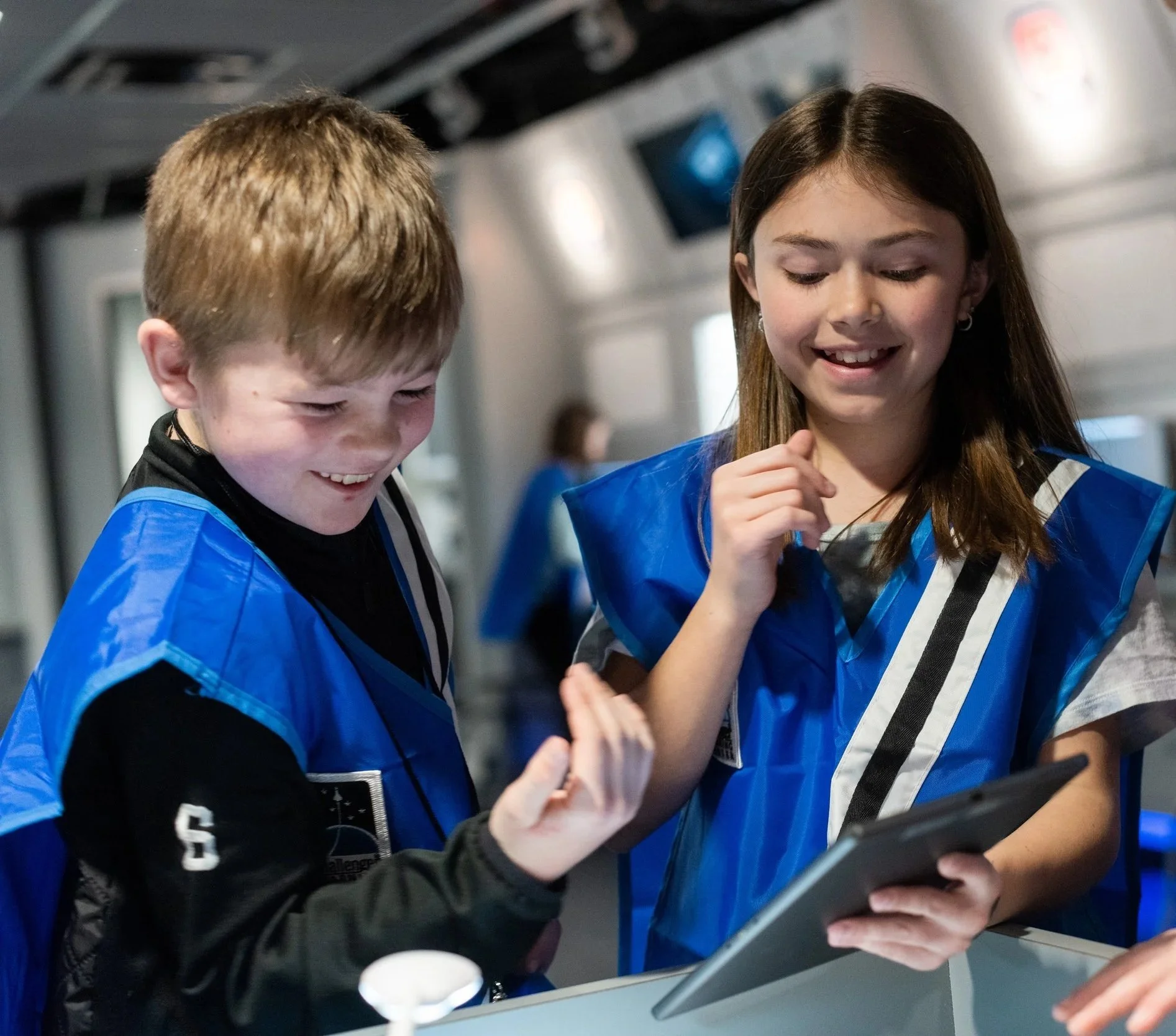 Two students wearing blue vests, smiling and looking at an ipad
