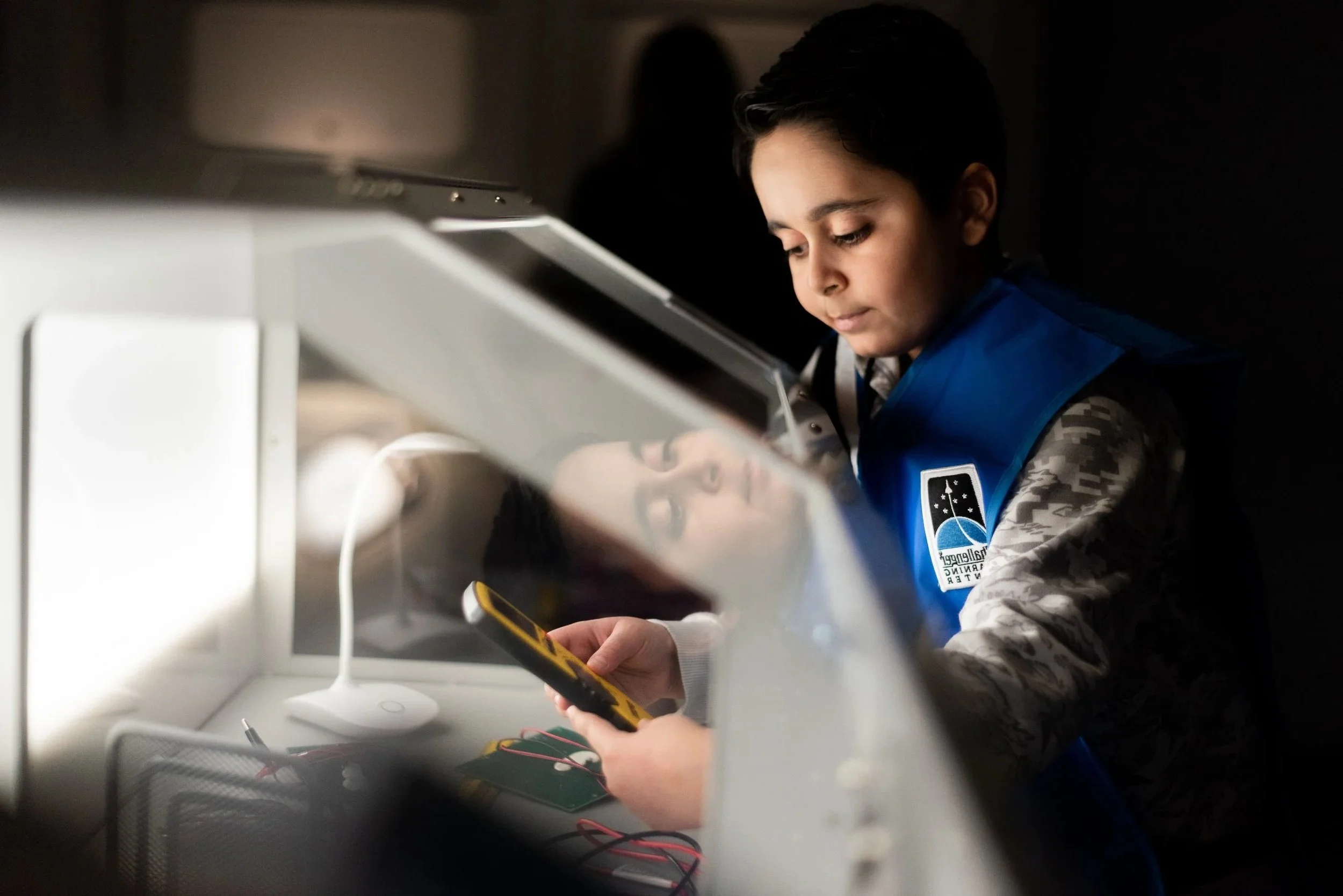 A young student wearing a blue NASA vest works with electronic equipment, with their reflection visible in a screen or monitor.