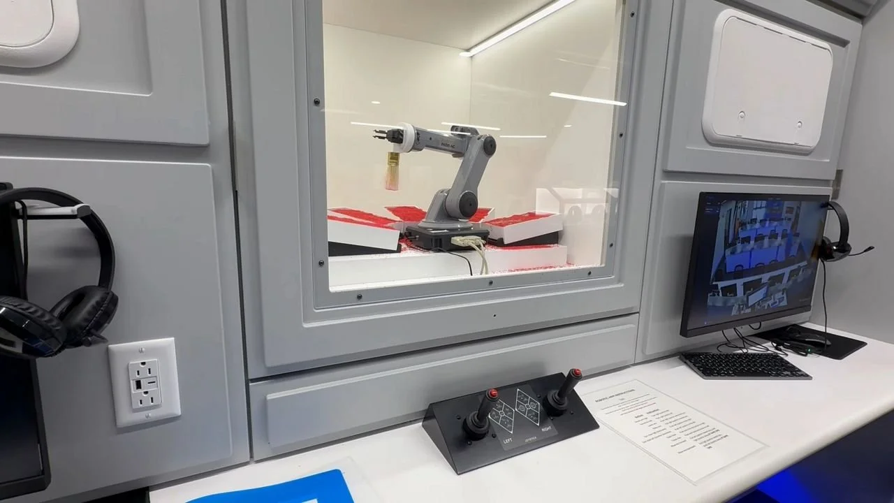 Laboratory robotic arm inside a glass-walled enclosure with a computer monitor and control joysticks on the desk outside.
