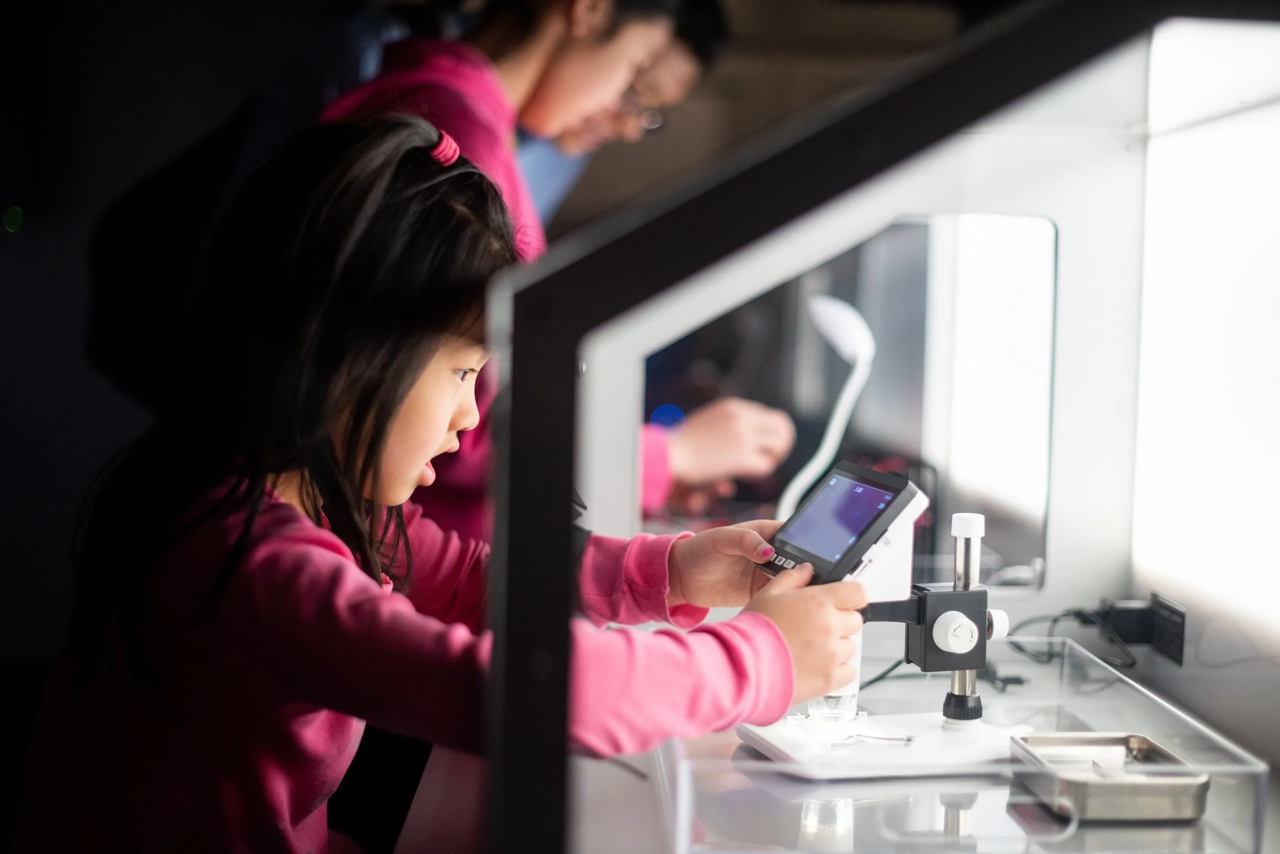Young girl in pink sweatshirt conducting a science experiment with a microscope, digital measuring device, and test tubes in a Challenger Center.