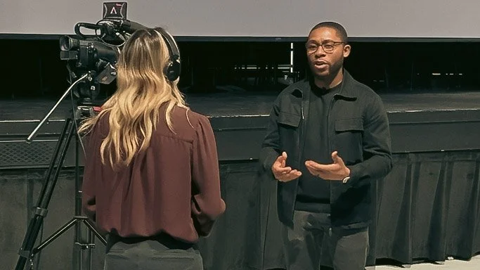 A woman with blonde hair and a maroon blouse is filming a man with glasses, wearing a black jacket, as he talks and gestures during an interview or presentation on a stage.