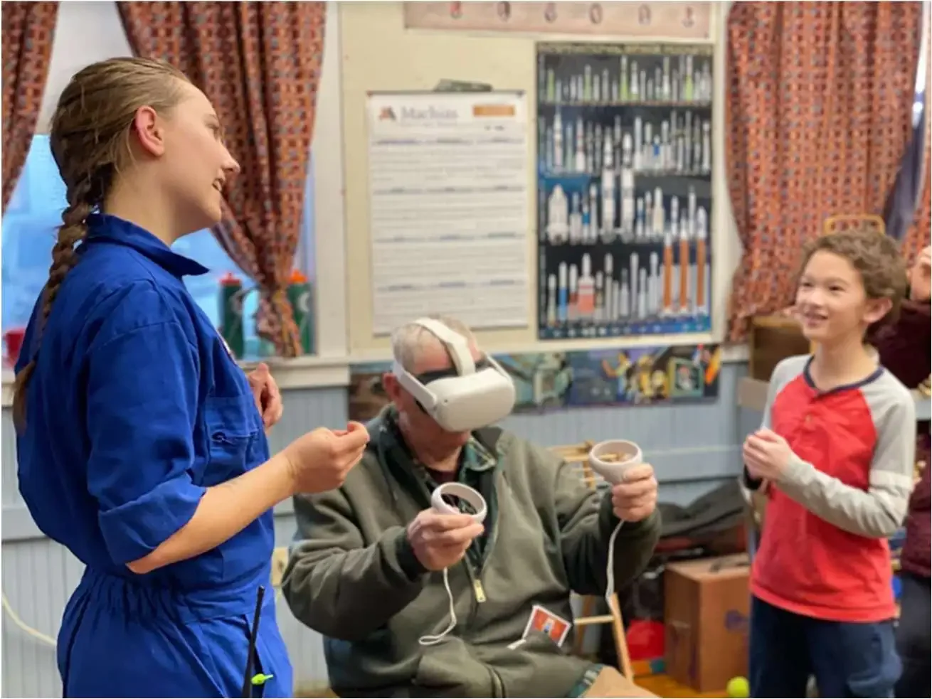 A young girl in a blue jumpsuit and a young boy in a red and gray shirt stand smiling and talking to an older man wearing a virtual reality headset and holding game controllers inside a room with colorful curtains and framed posters on the wall.